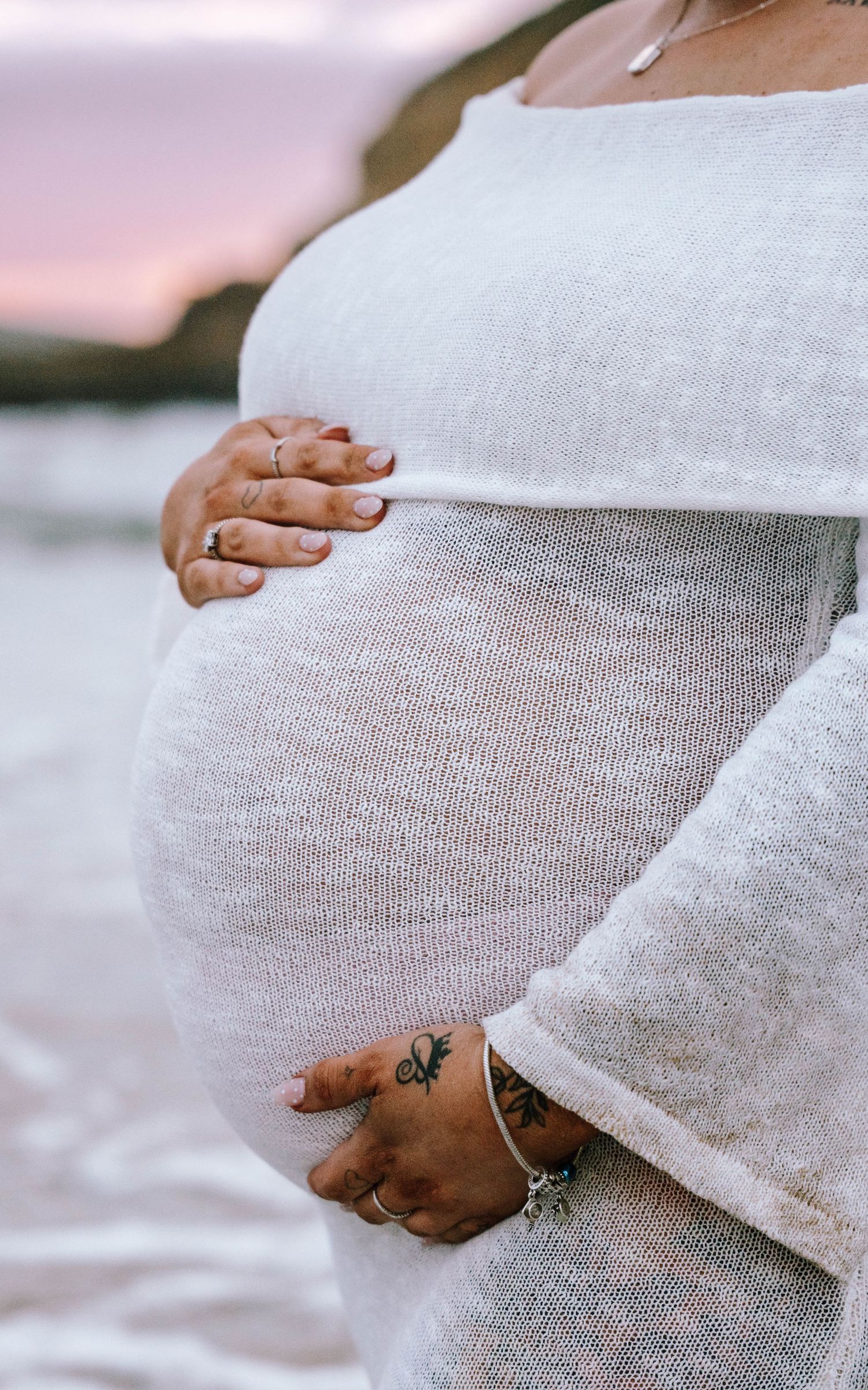 Close-up of pregnant belly in white dress with hands gently cradling the bump, showing small tattoo and bracelet.