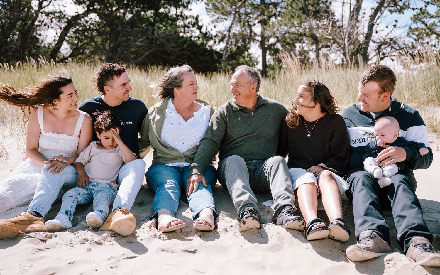 Multi-generational family laughing together on a sandy beach, seated in a row on a sunny day.