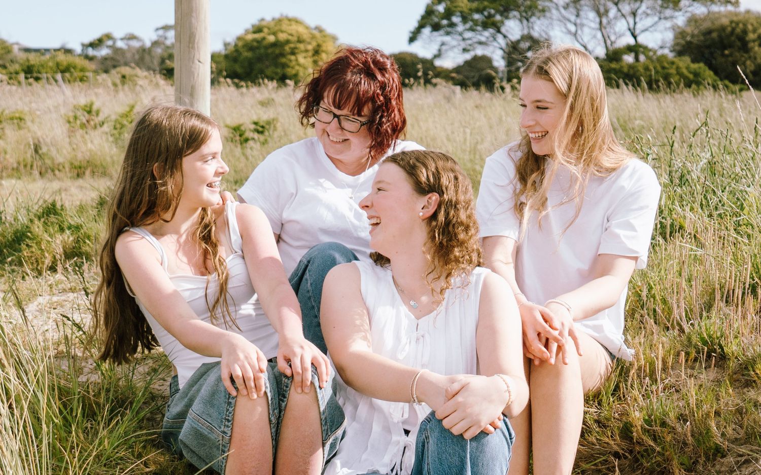 Four women sitting together laughing in a sunny grassy field, wearing white and light blue casual outfits.