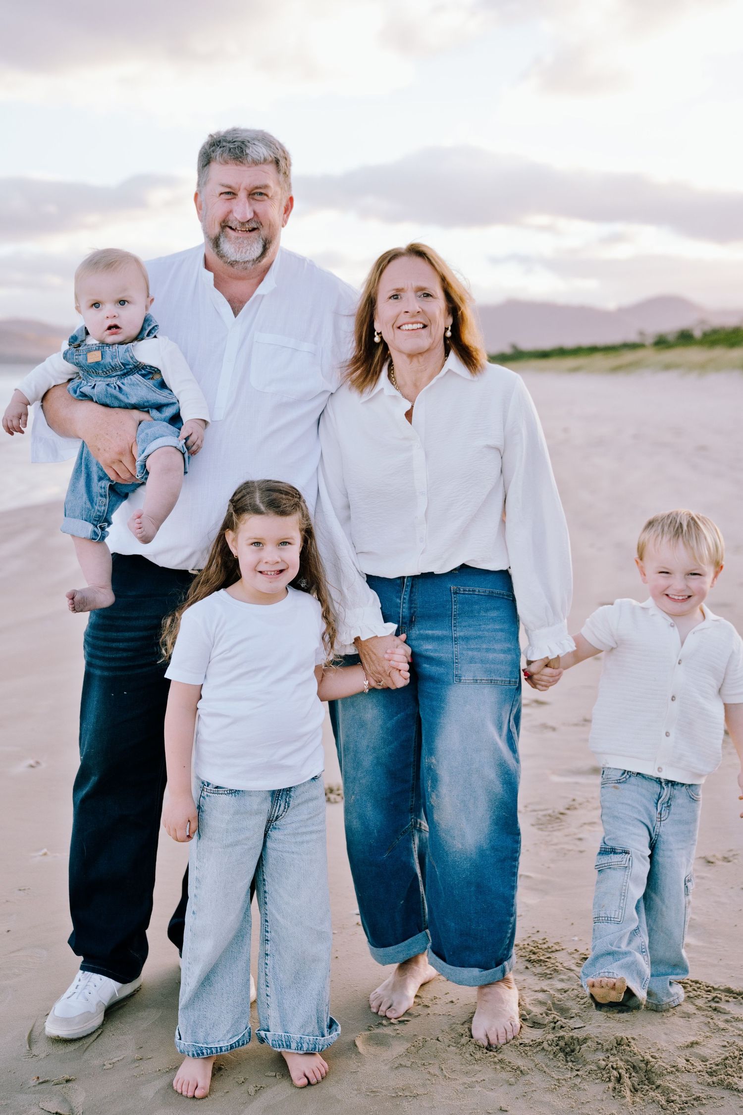Happy family of five posing on sandy beach at sunset, parents in white shirts and jeans with three young children.