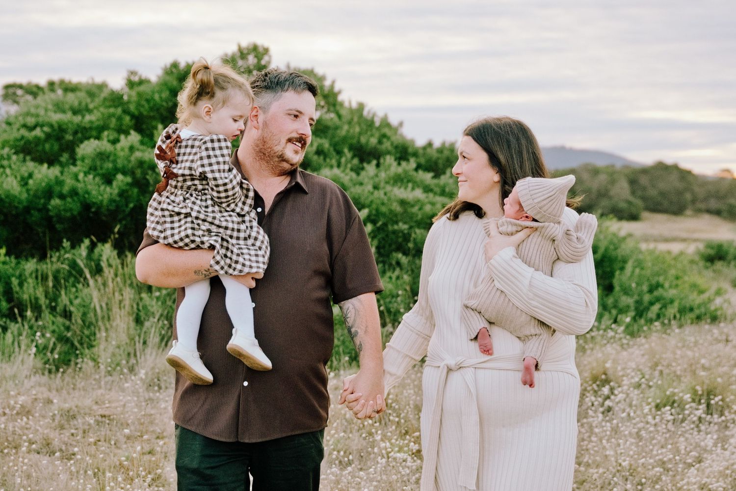 A family of four poses outdoors in a field, parents holding two young children, smiling warmly in natural light.