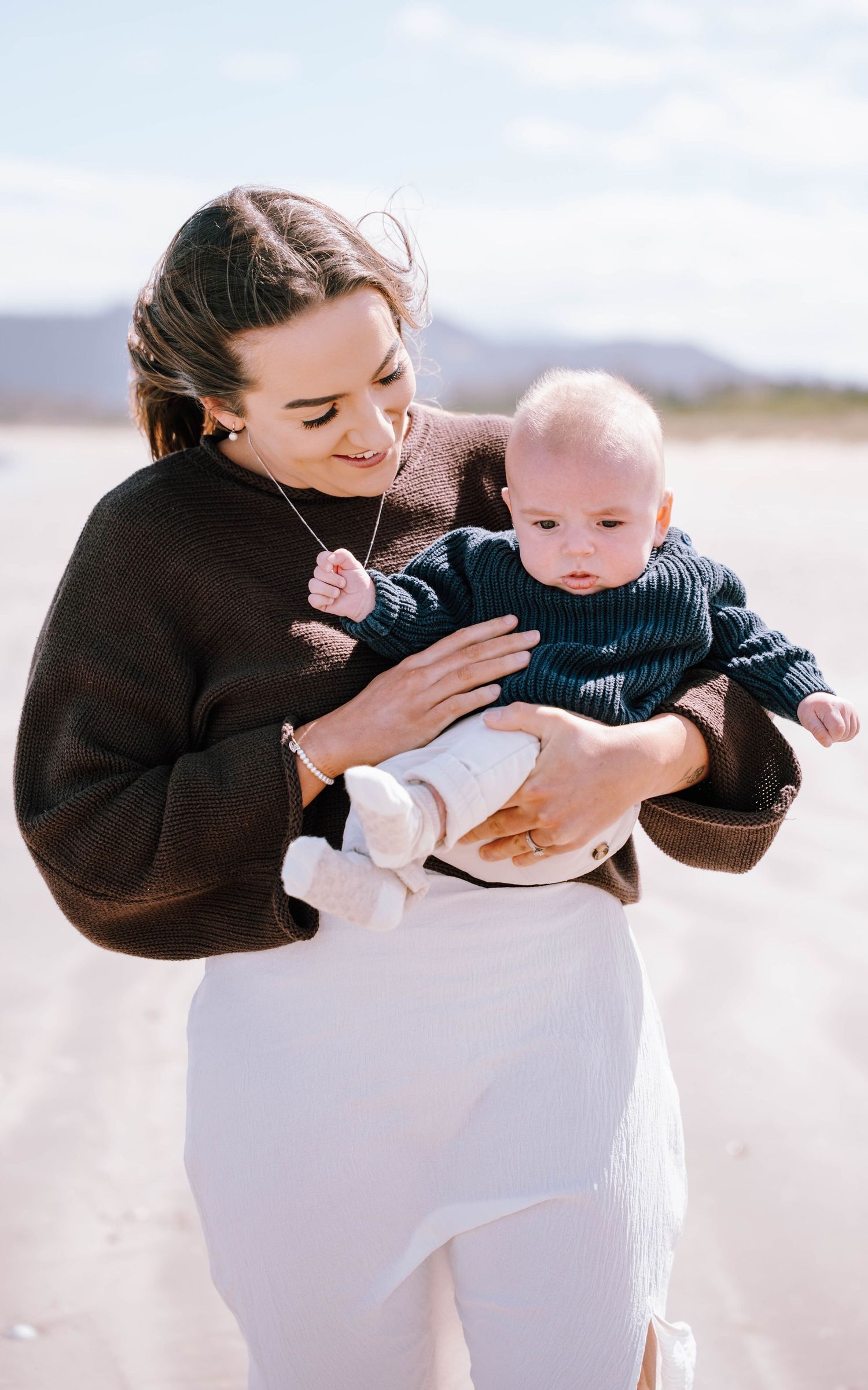 A parent in a brown sweater and white pants holds a baby wearing a navy striped outfit on a bright sunny day.