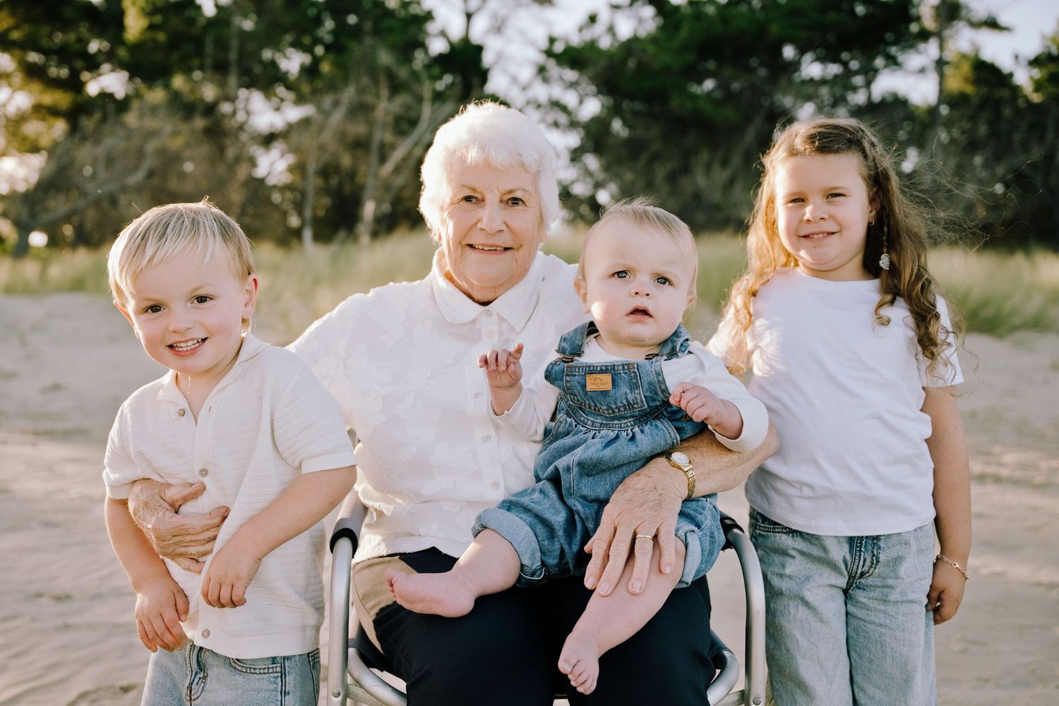 Elderly grandmother sitting on beach sand smiling with three young grandchildren in matching white shirts and jeans.
