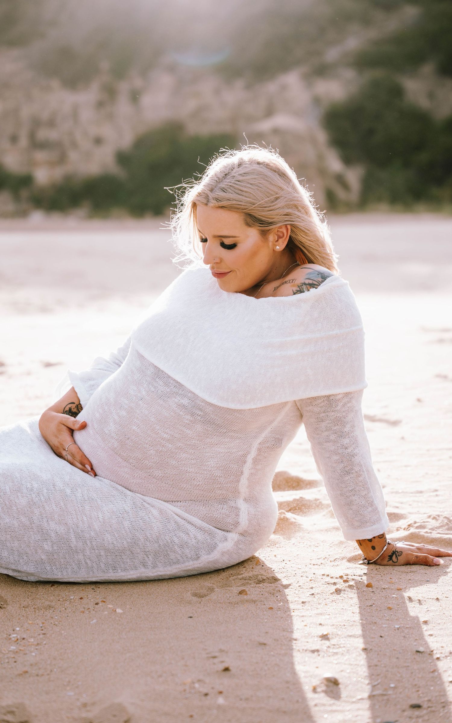 Woman in white lace dress poses gracefully on sandy beach at sunset creating ethereal maternity photos.