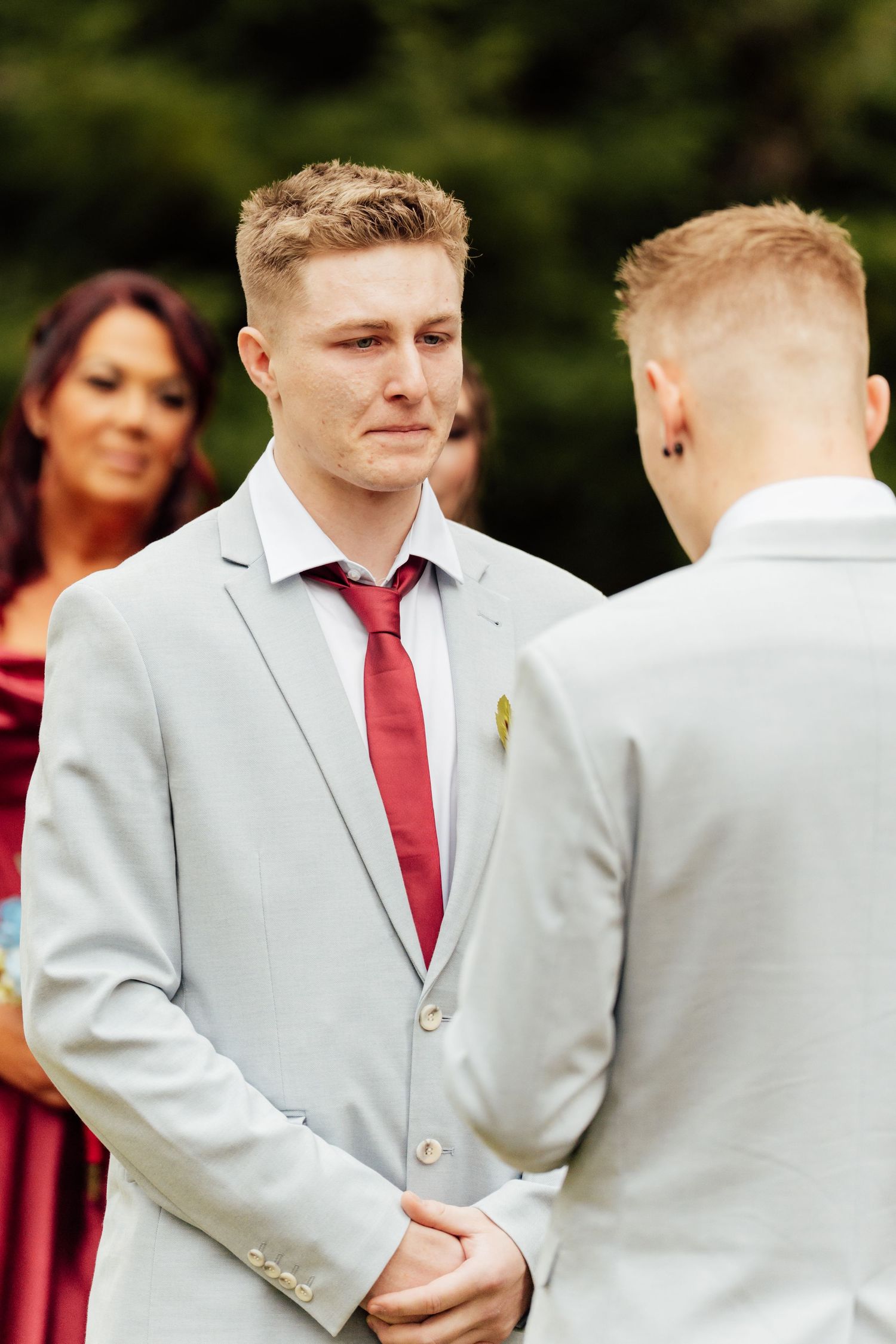Two grooms in gray suits with red ties exchange vows at an outdoor wedding ceremony.