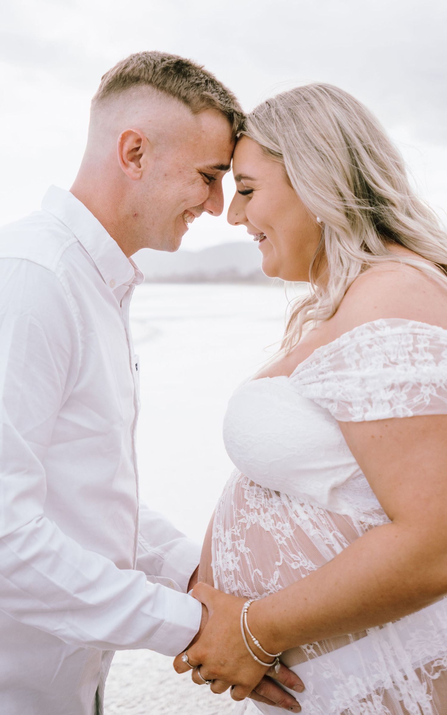 An intimate maternity portrait showing a couple in white sharing a tender moment against an overcast sky.
