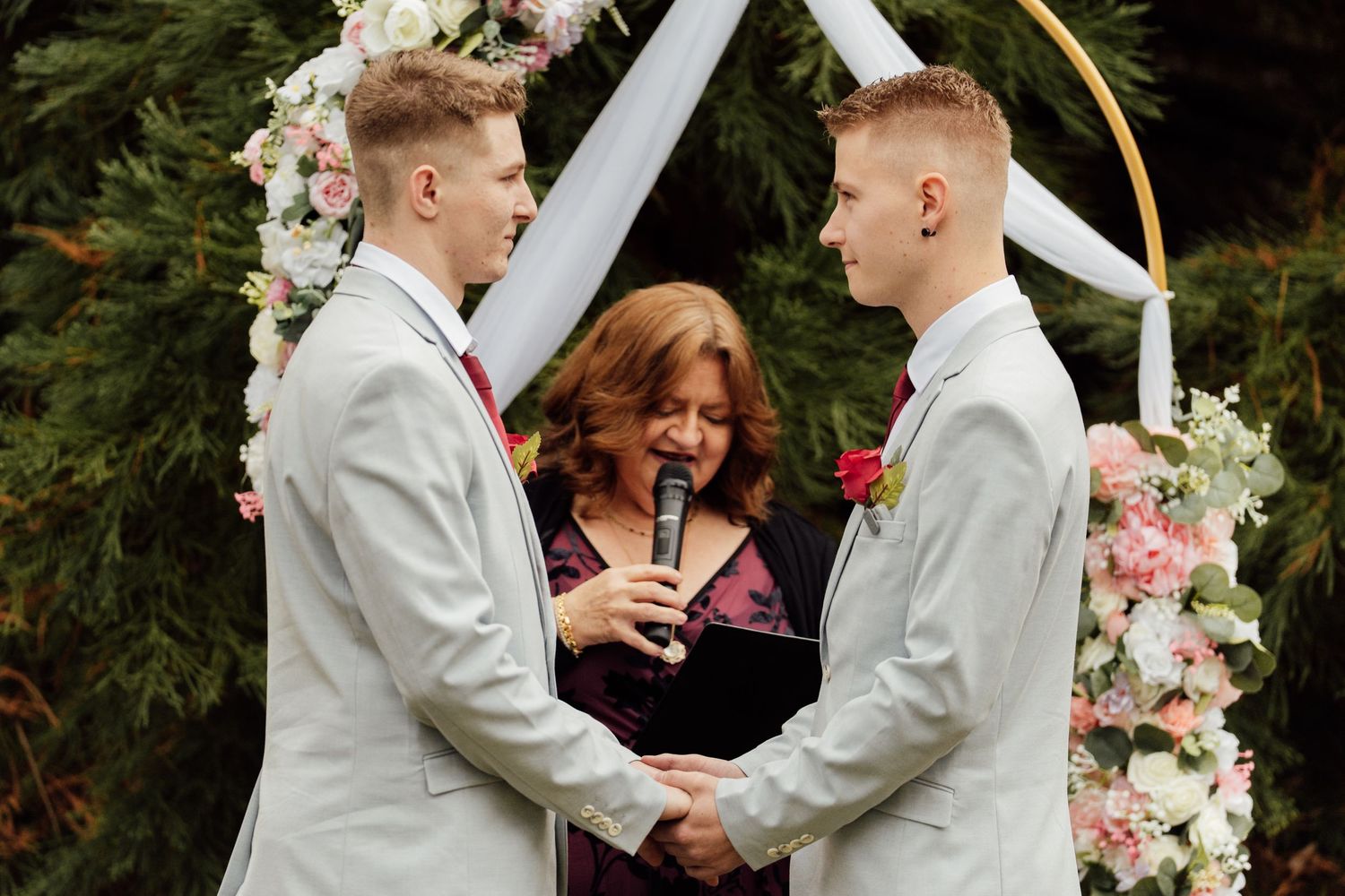 Two grooms in gray suits exchange vows at an outdoor wedding ceremony with a floral arch backdrop.
