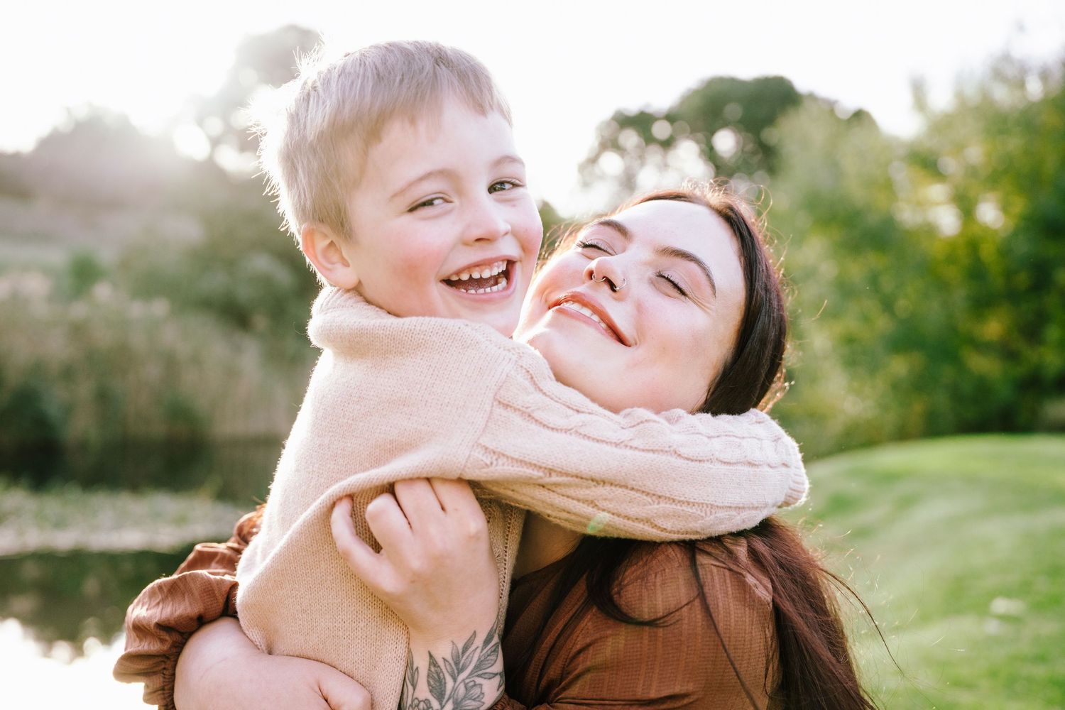 A joyful mother and young son share a warm embrace outdoors, laughing together near a sunlit pond.