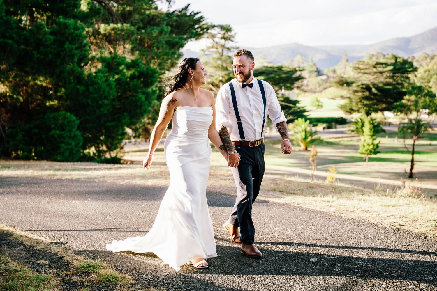 A bride and groom walk hand in hand down a sunlit path surrounded by lush greenery on their wedding day.