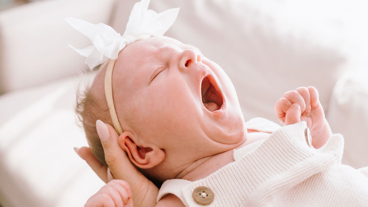 Newborn baby in white clothing yawns while lying on soft white bedding.