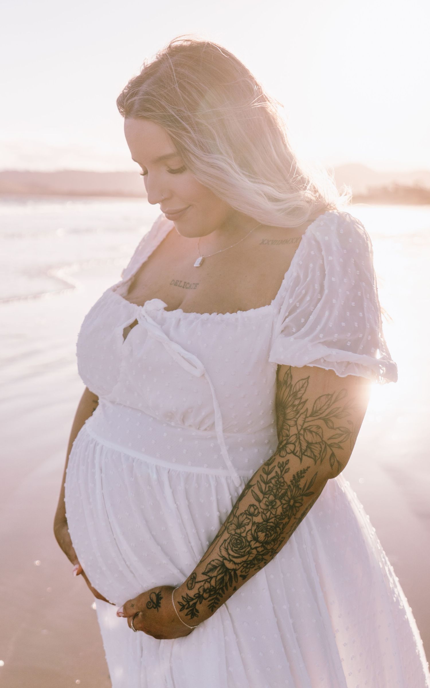 A pregnant woman in a white lace dress stands on a beach at sunset with sunlight creating a dreamy glow.