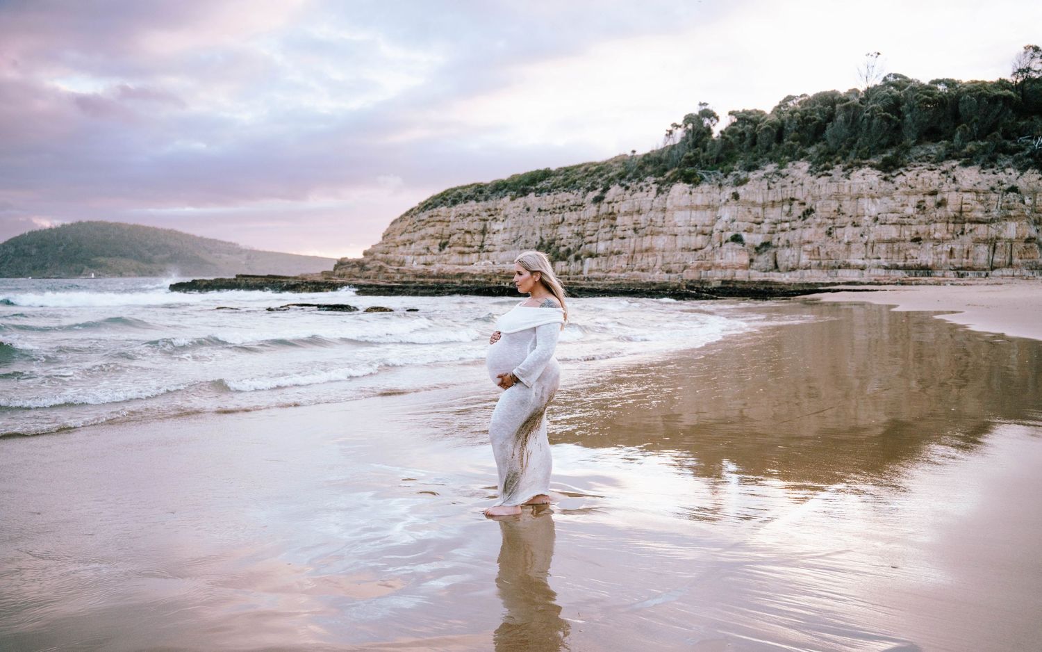 A person in white clothing walks along a scenic beach with dramatic limestone cliffs and crashing waves at sunset.