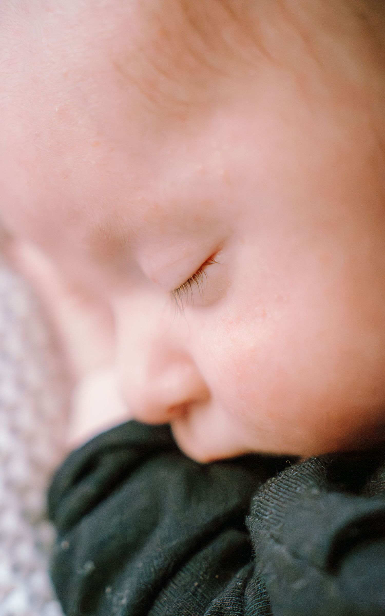 Close-up profile view of a sleeping baby in a dark green outfit, showing delicate eyelashes and soft skin.