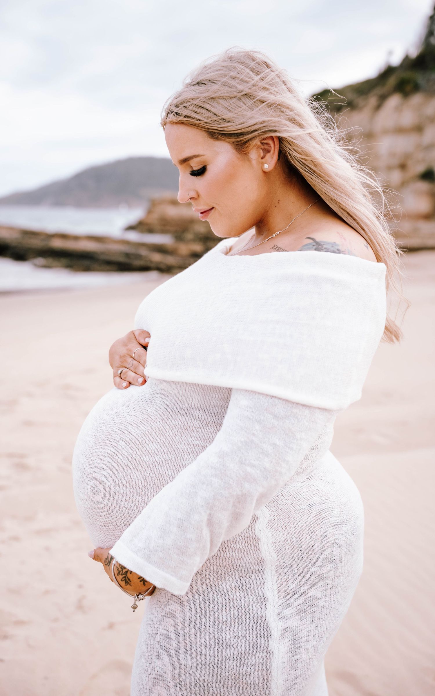 A pregnant woman in a white sweater poses thoughtfully on a beach with cliffs and ocean in the background.