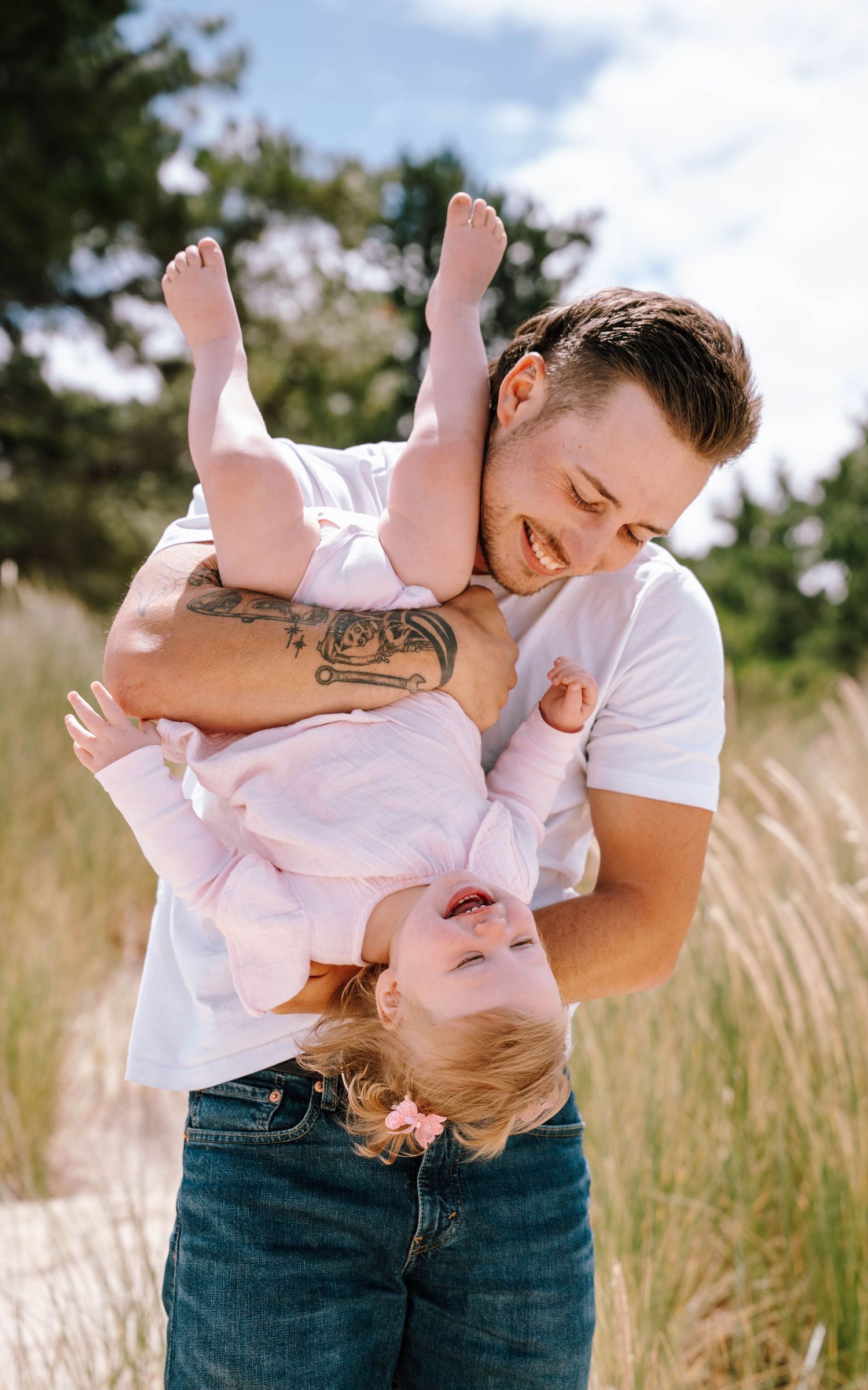 Parent in white shirt and jeans playfully holds and kisses small child wearing pink dress in sunny outdoor field.