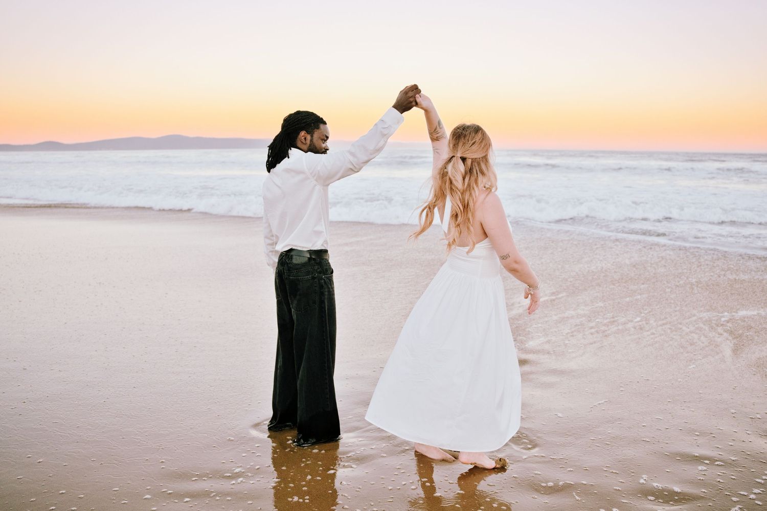 Couple dancing barefoot on a beach at sunset, bride in white dress, groom in dark pants, holding hands.
