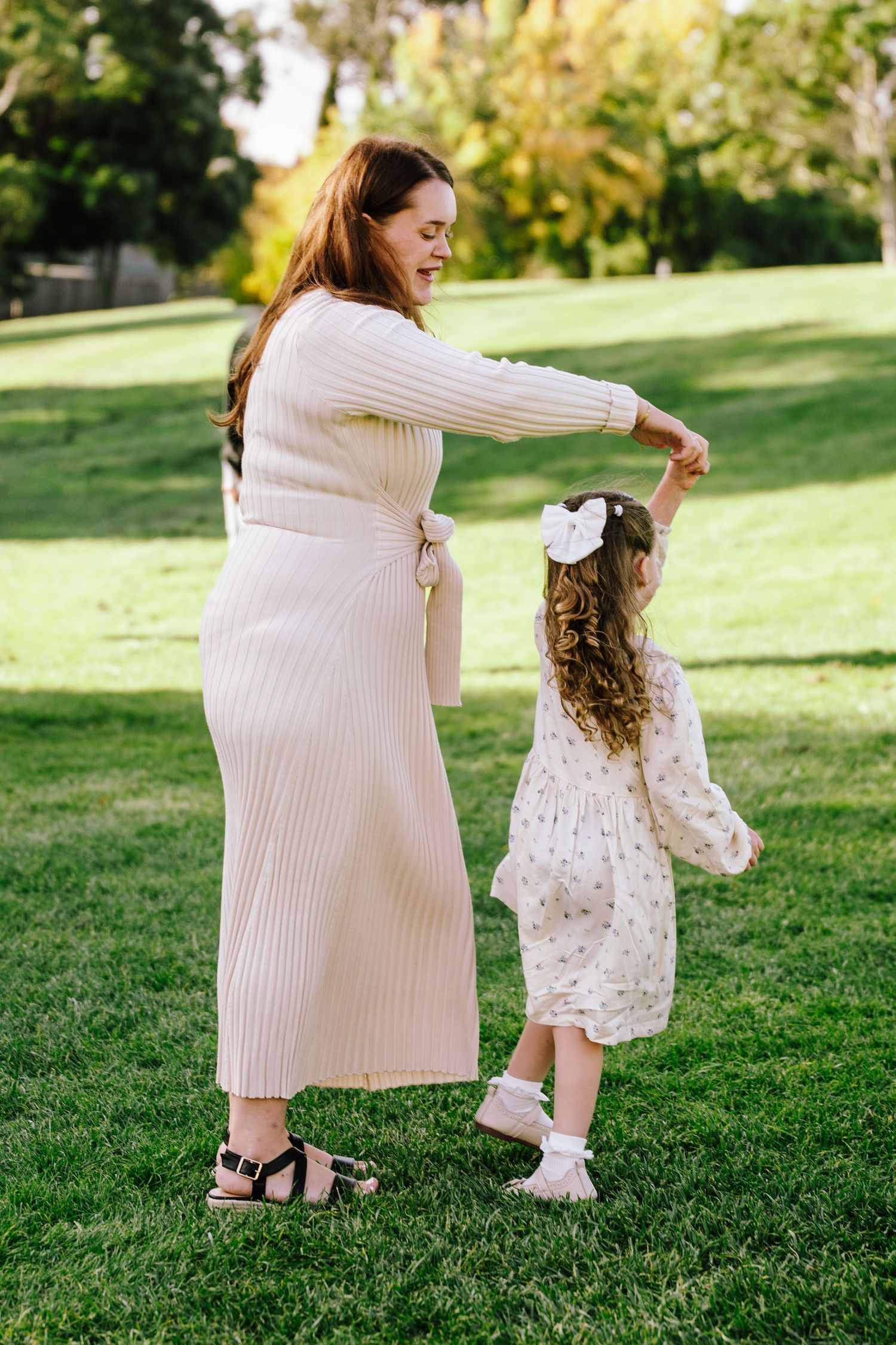 A mother in a cream knit dress twirls her young daughter in a floral dress on a green lawn in a sunny park.