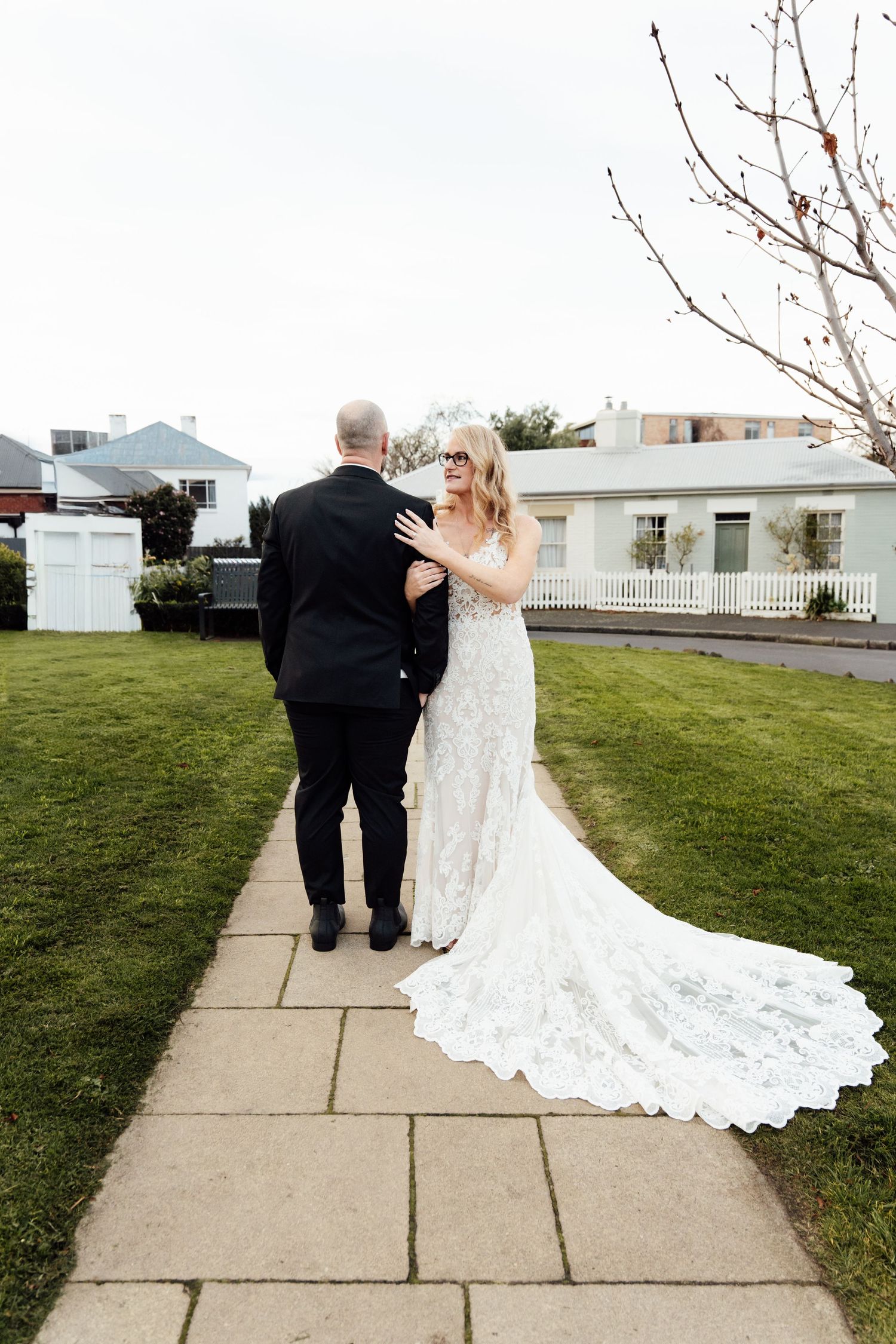 Bride in lace gown with train stands with groom on garden path during outdoor wedding ceremony.