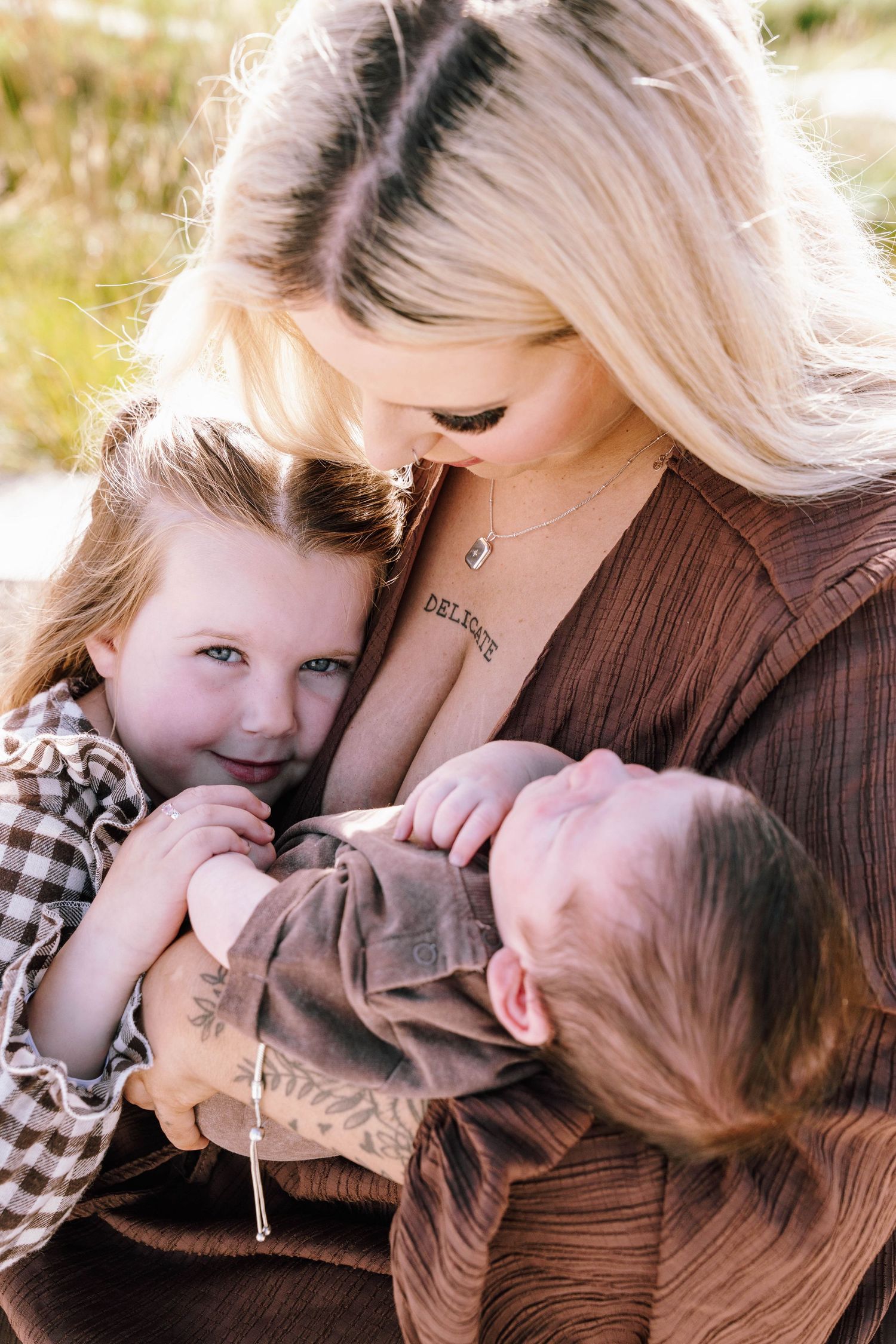 A blonde mother tenderly holds her newborn and young daughter outdoors in a warm family portrait.