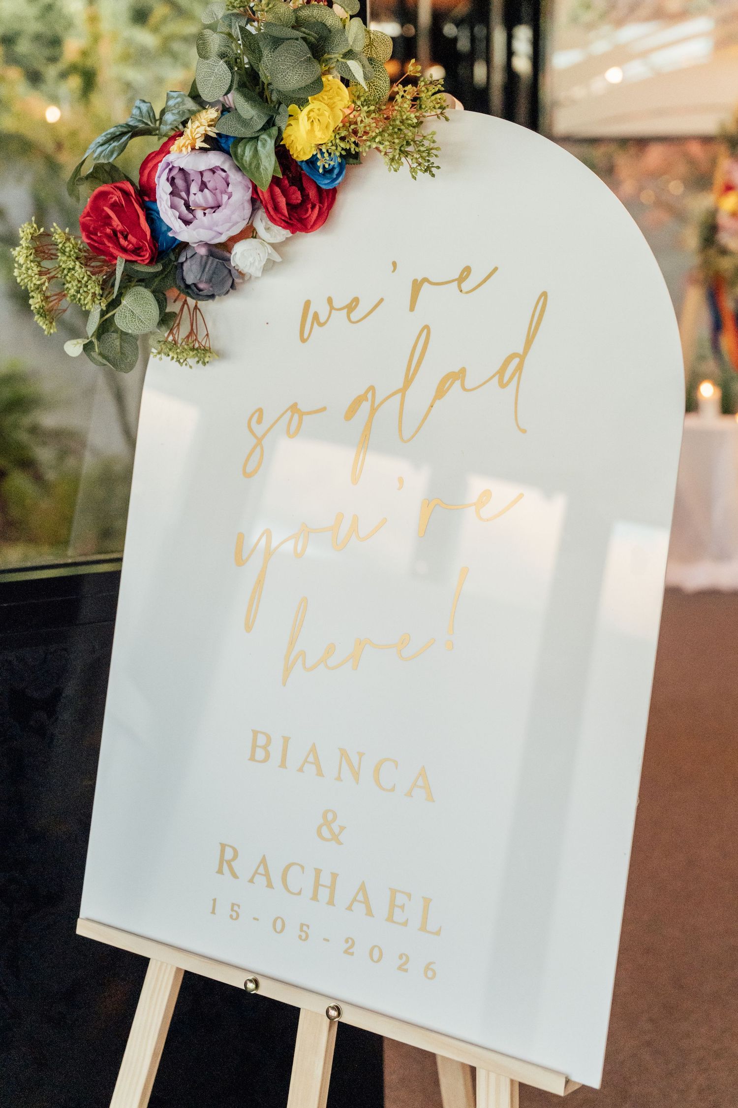 Wedding welcome sign for Bianca & Rachael on 15-08-2025, with gold script and colorful floral arrangement on easel.
