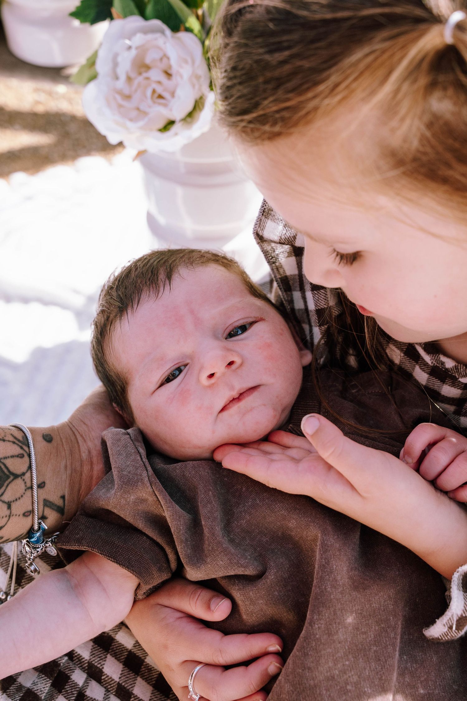 Older child gazing lovingly at a newborn baby wrapped in a brown blanket, held by an adult outdoors.