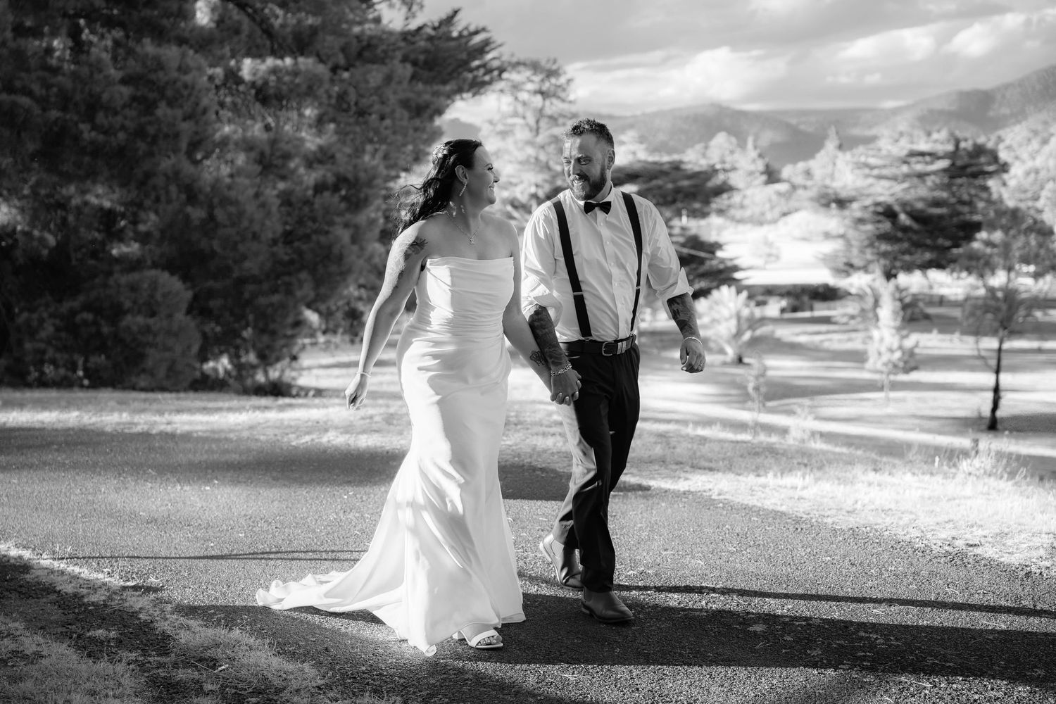 Black and white photo of a bride and groom walking hand in hand on a path surrounded by trees on their wedding day.