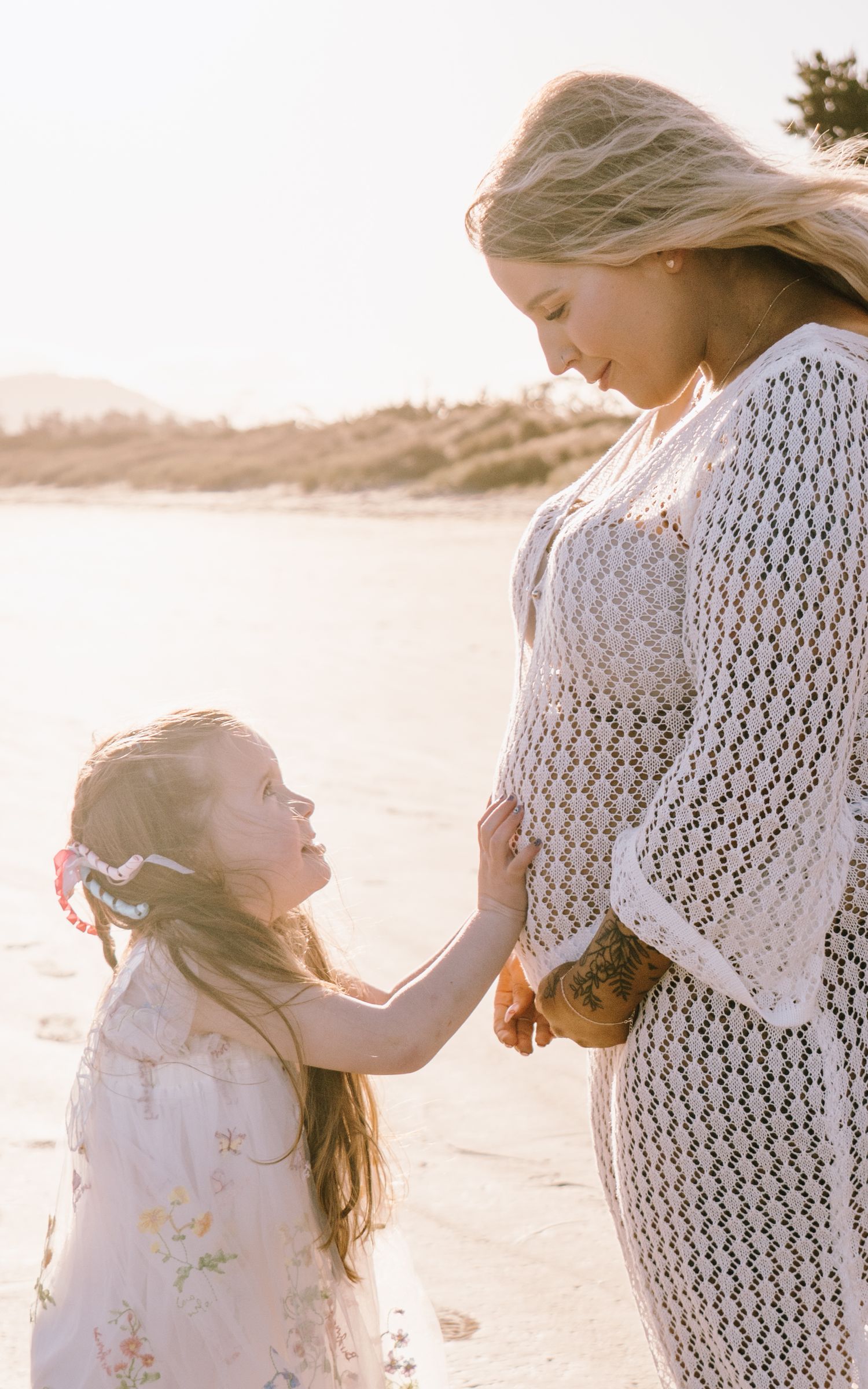 A mother and daughter in white lace dresses embrace on a sunny beach at sunset.