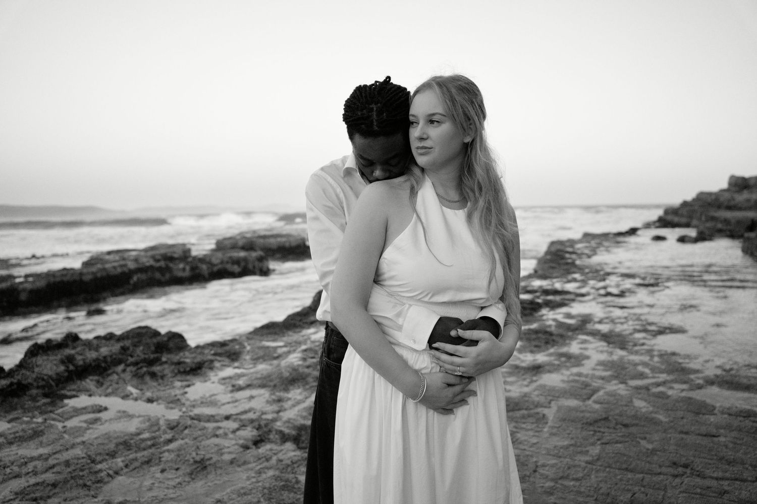 Black and white photo of an interracial couple embracing on a rocky beach, woman in white dress holding a phone.