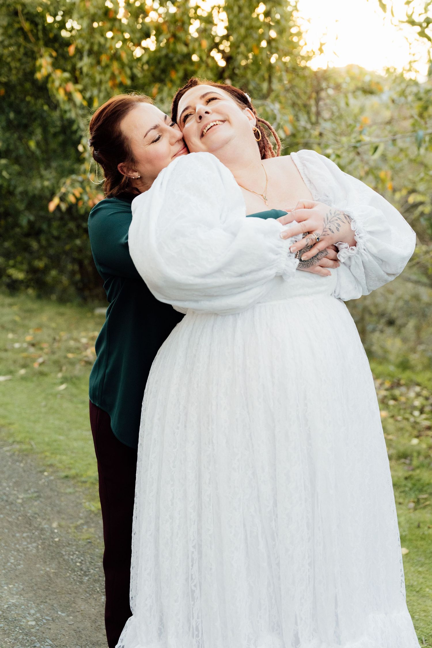 Two women embrace outdoors, one in a white lace bridal gown with puff sleeves, the other in a dark green dress.