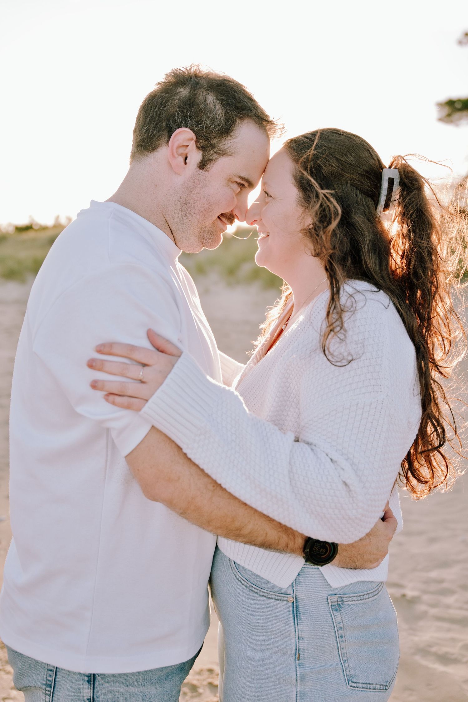 Romantic couple embracing face-to-face on a sandy beach at golden hour, woman in white sweater and jeans.