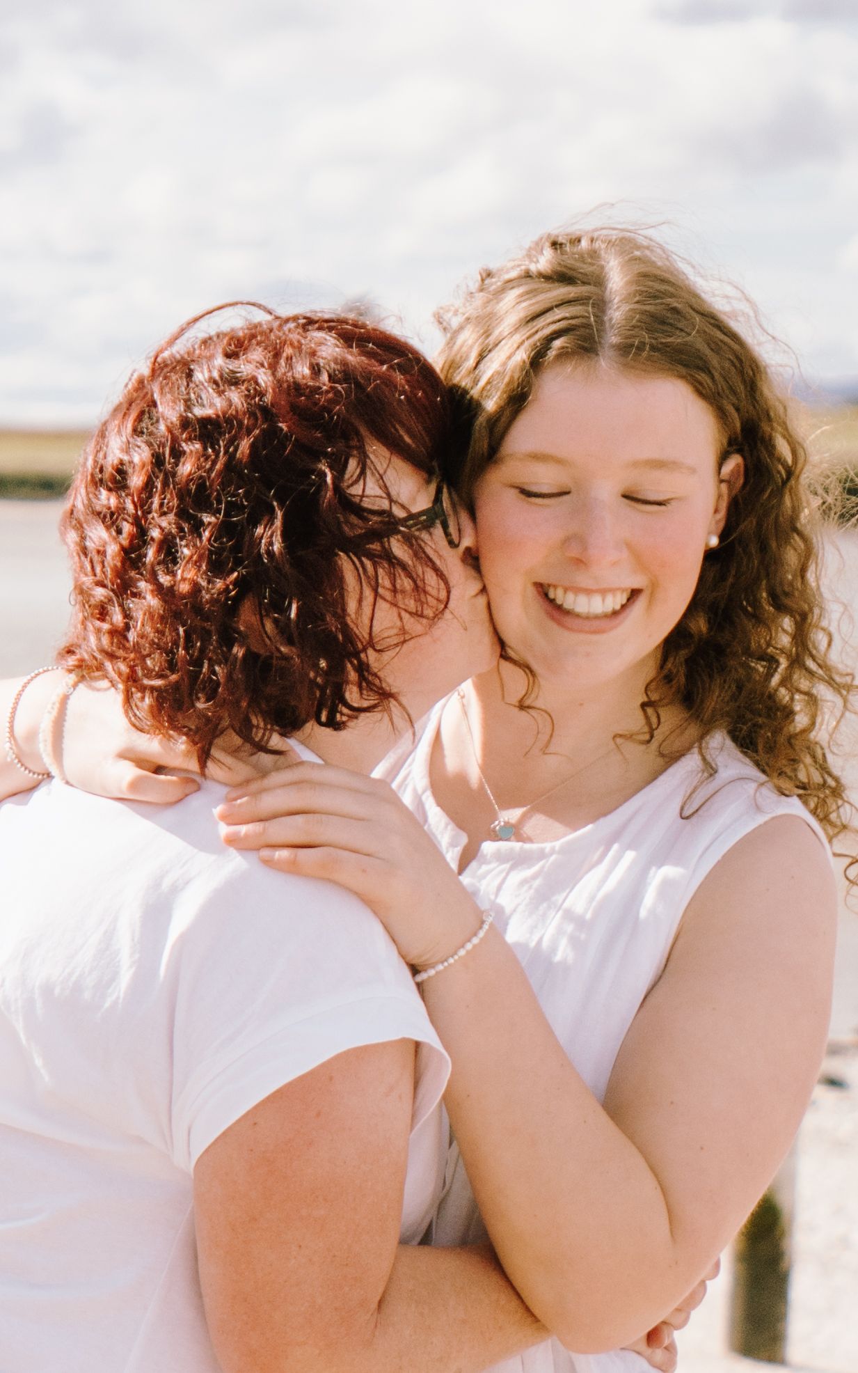 Two women in white tops embracing and smiling outdoors near water on a sunny day.