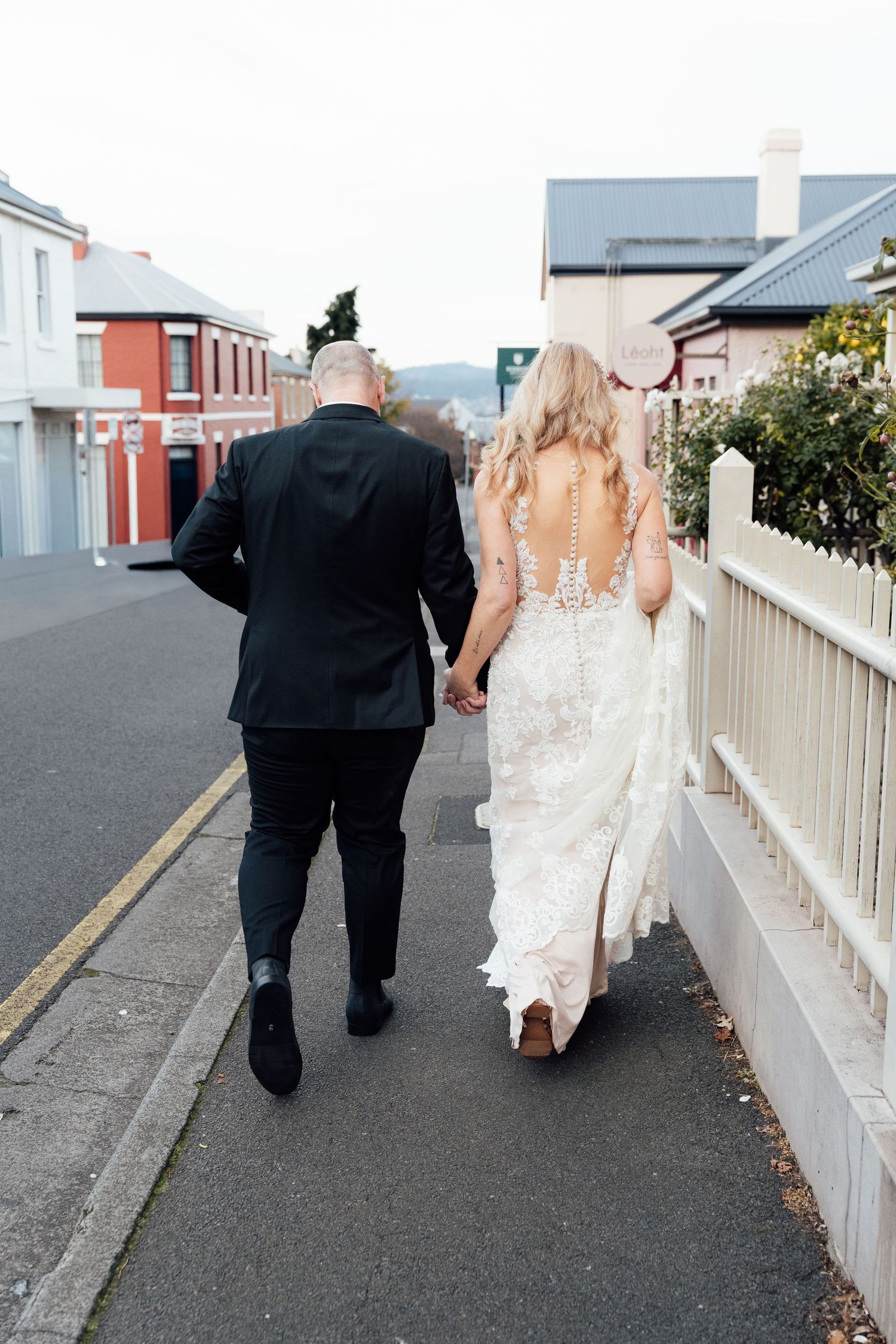 Bride and groom walking hand in hand down a street, bride wearing a backless lace wedding gown.