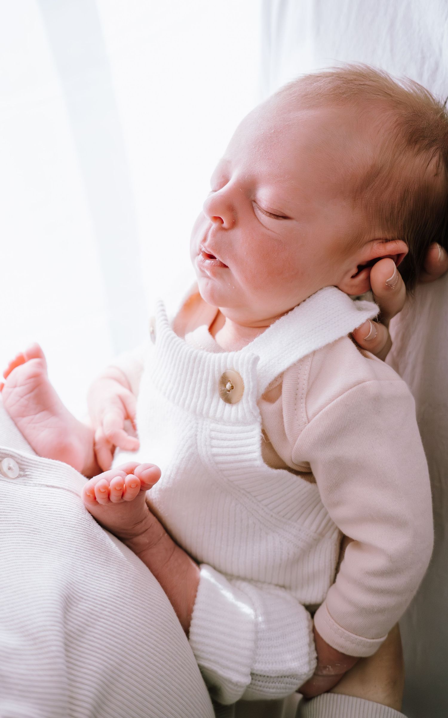 A sleeping newborn baby in white clothing rests peacefully against a light background.