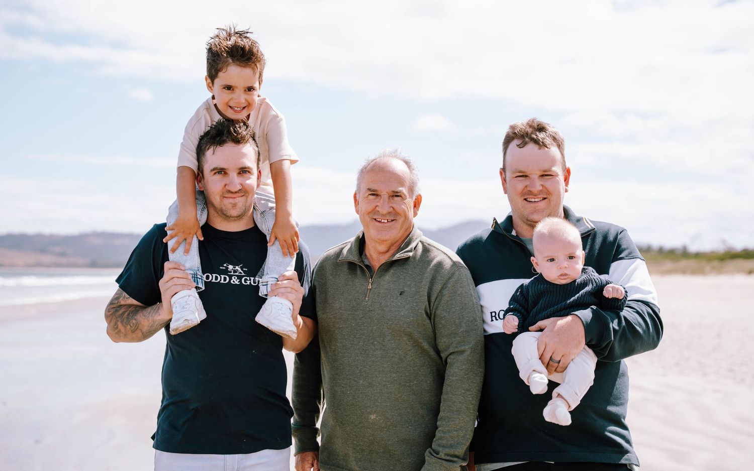 Three generations of men smile on a beach, with a child on shoulders and a baby in arms.