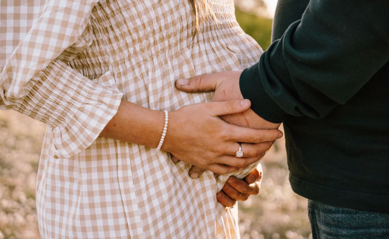Close-up of hands wearing rings holding each other tenderly against a gingham pattern dress outdoors.