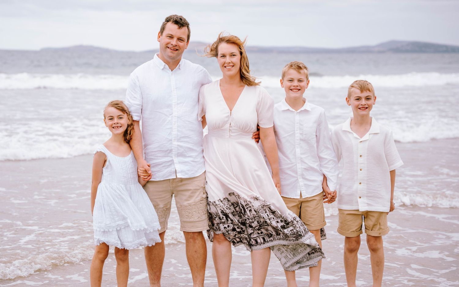 A family dressed in white and beige outfits poses together on a sandy beach with ocean waves in the background.