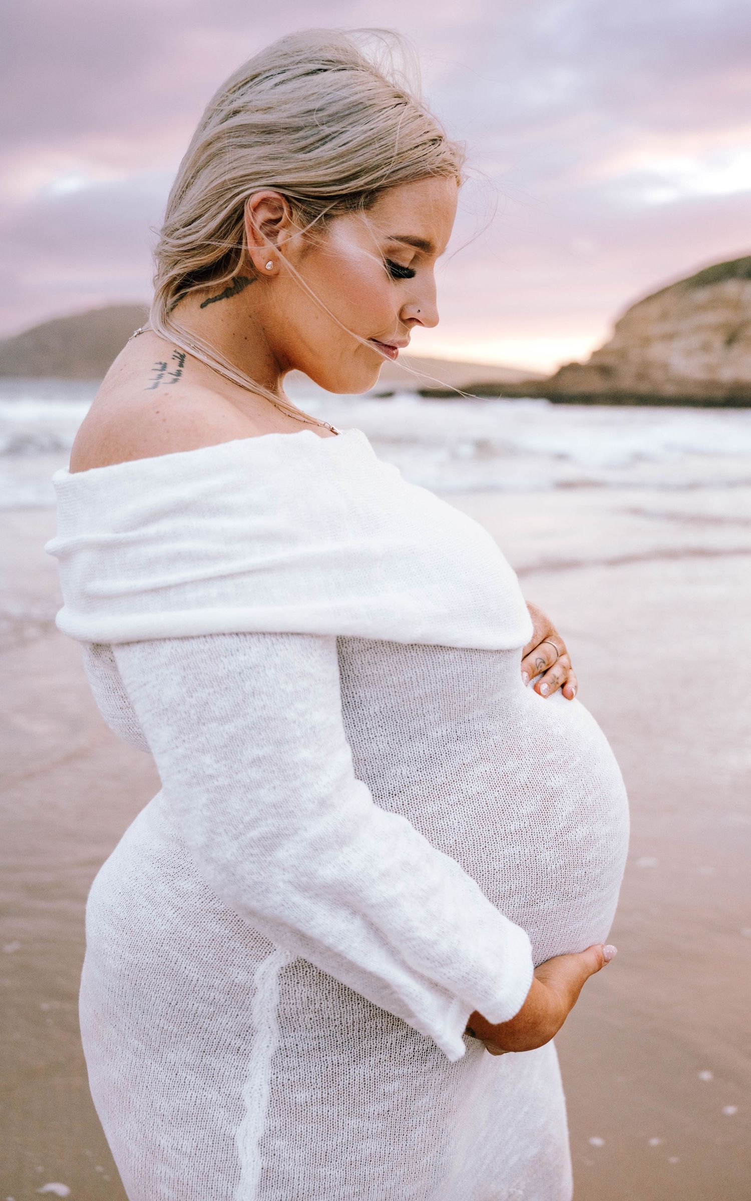 Expectant mother in white sweater poses on beach at sunset holding her pregnant belly.