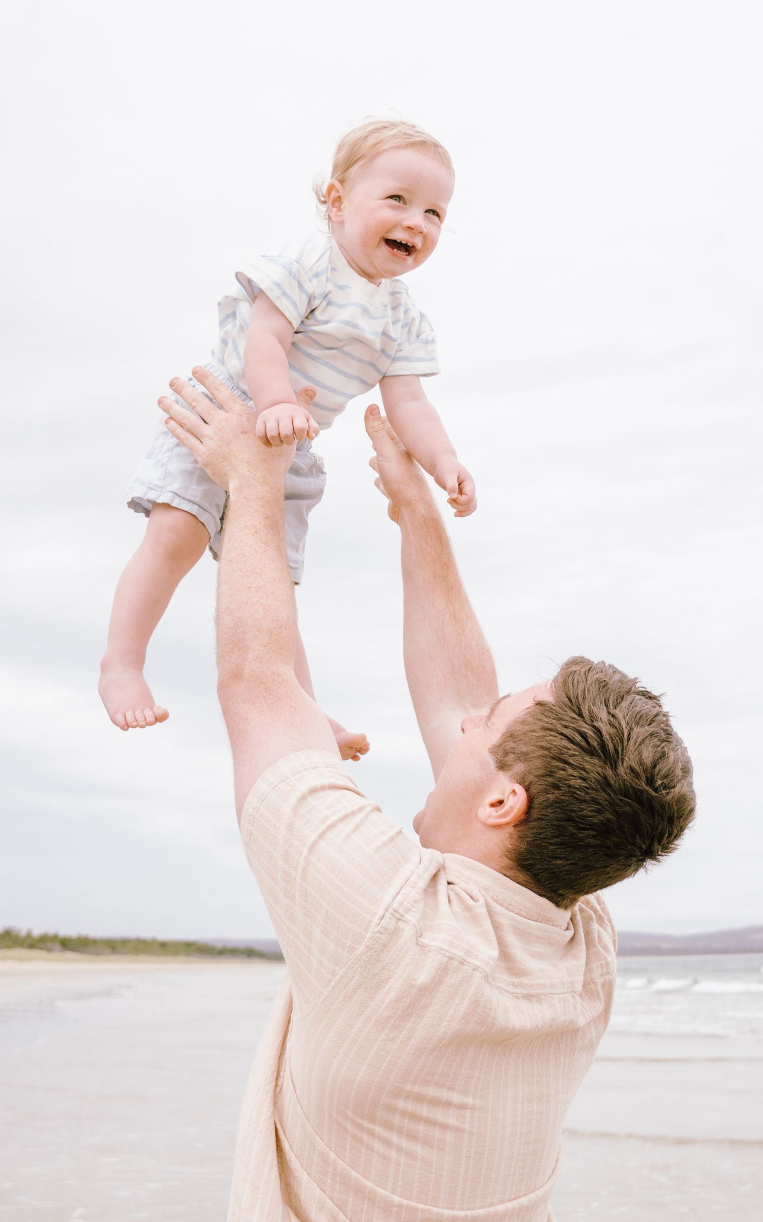 A joyful baby is lifted high into the air at the beach while wearing light colored clothing.