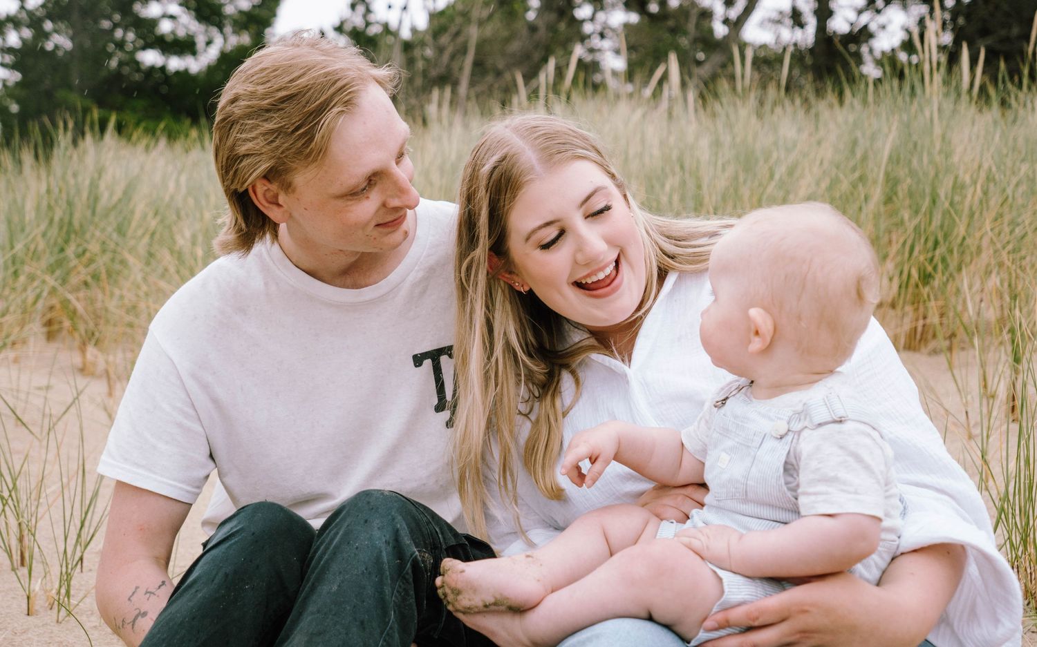 A loving family shares tender moments together while sitting in tall grass during a casual outdoor photo session.