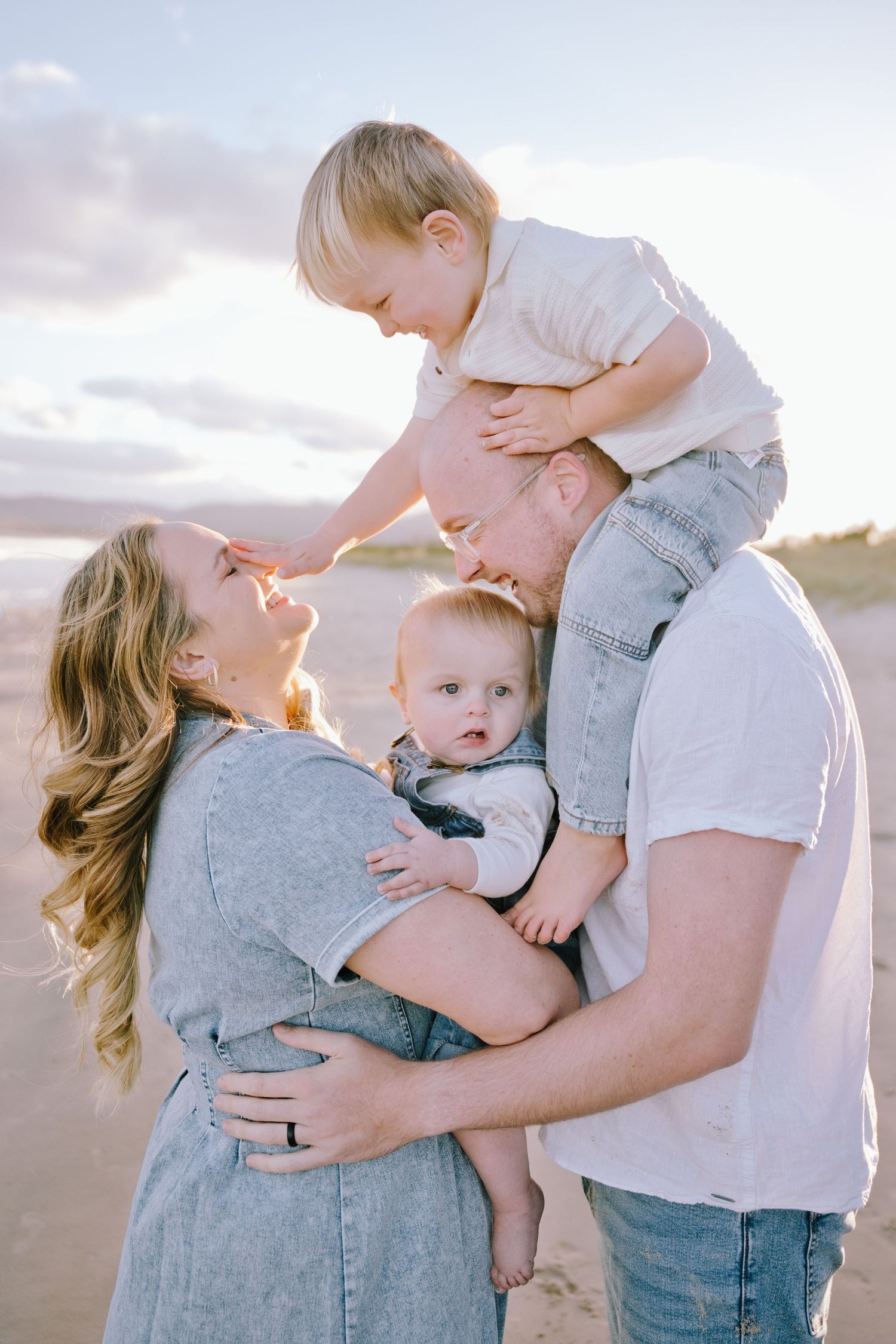 Family of four enjoying a beach sunset, mom and dad holding two young children, smiling and laughing together.