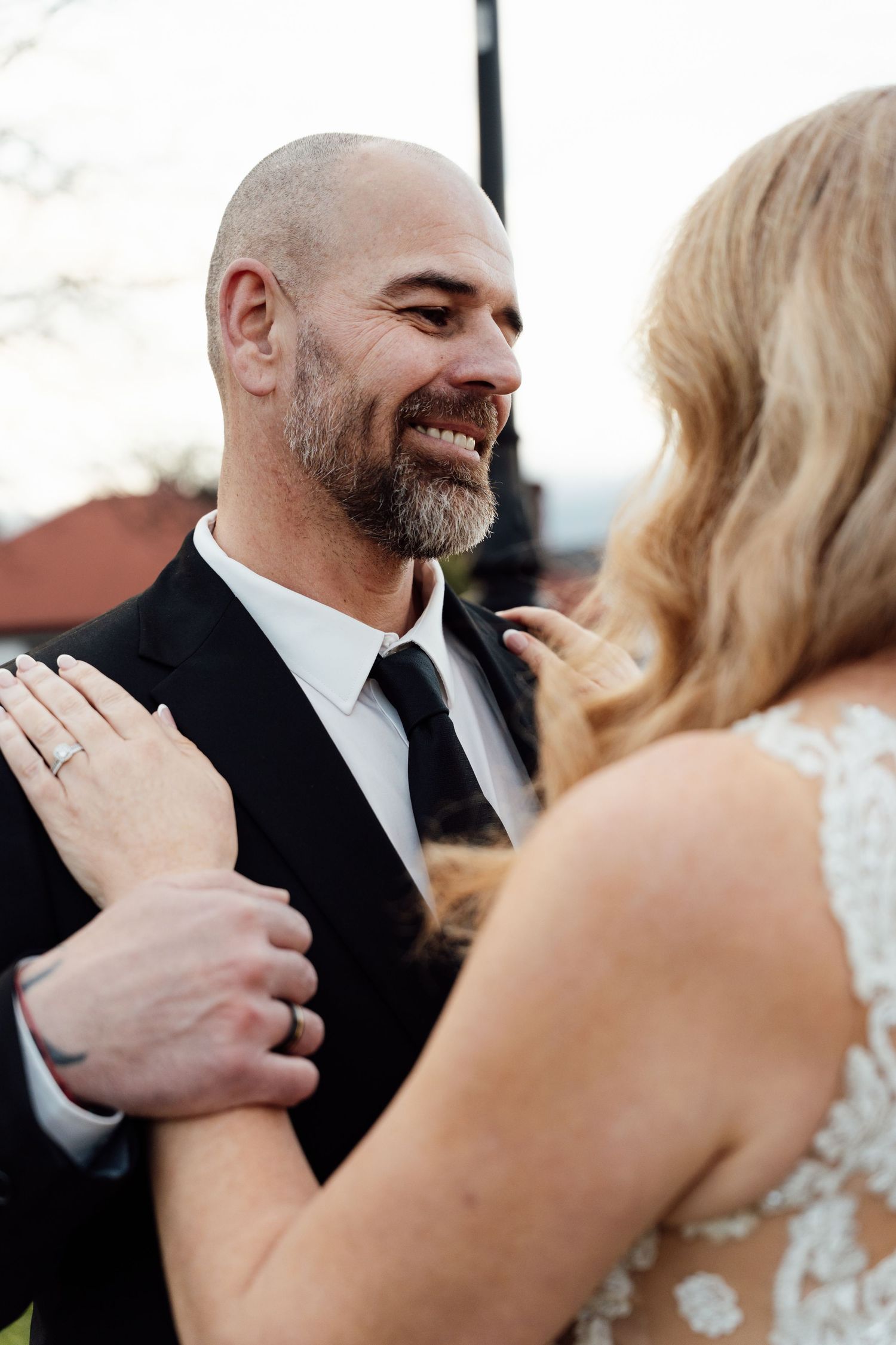Smiling bald groom with beard in black suit embracing blonde bride during outdoor wedding ceremony.