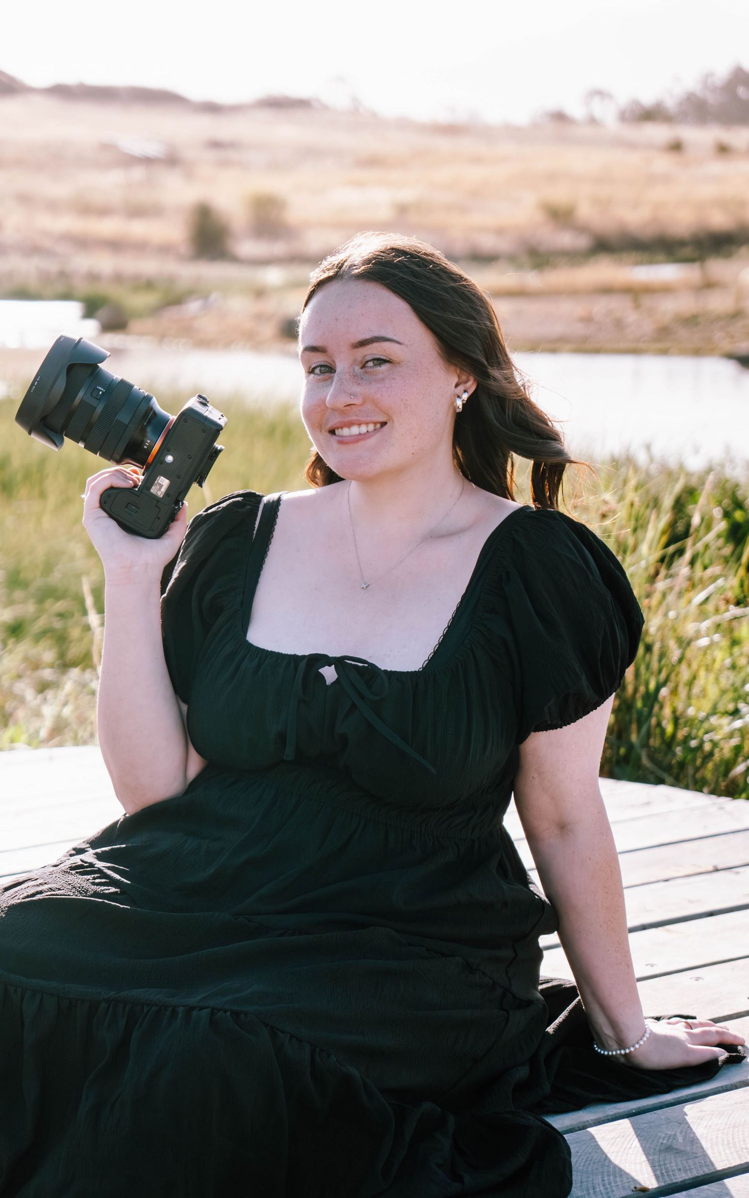 A person in a black dress poses outdoors on a wooden deck near a lake during a sunny day.