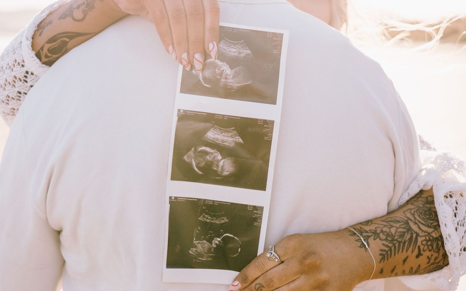 A person holds three ultrasound sonogram photos while wearing a white robe and displaying a tattoo on their hand.