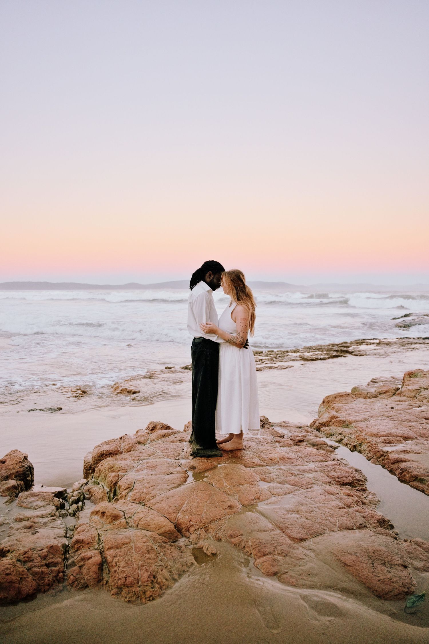 Couple embracing on rocky beach at sunset, woman in white dress, romantic ocean backdrop with pink sky.