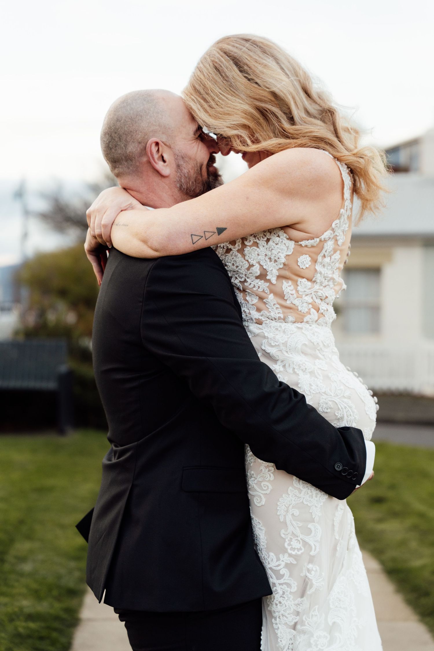 Bride and groom share an intimate embrace outdoors, with the bride in lace gown and groom in black suit.