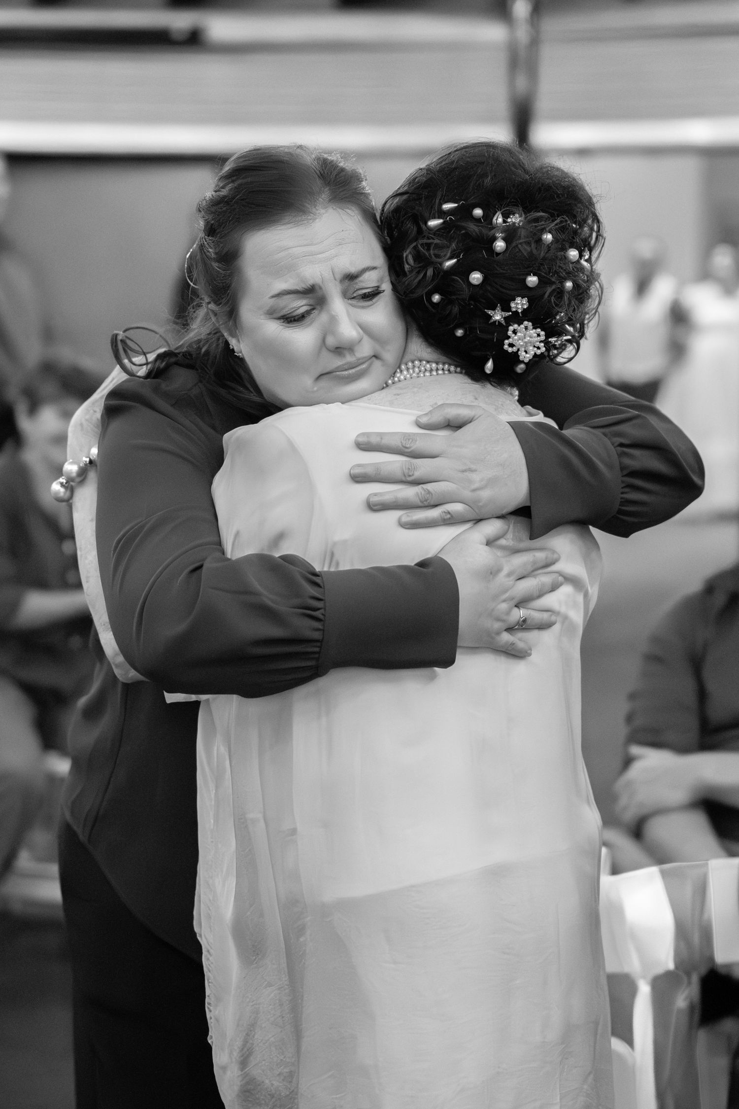 Black and white photo of two women sharing an emotional hug at a wedding ceremony, bride in white gown.