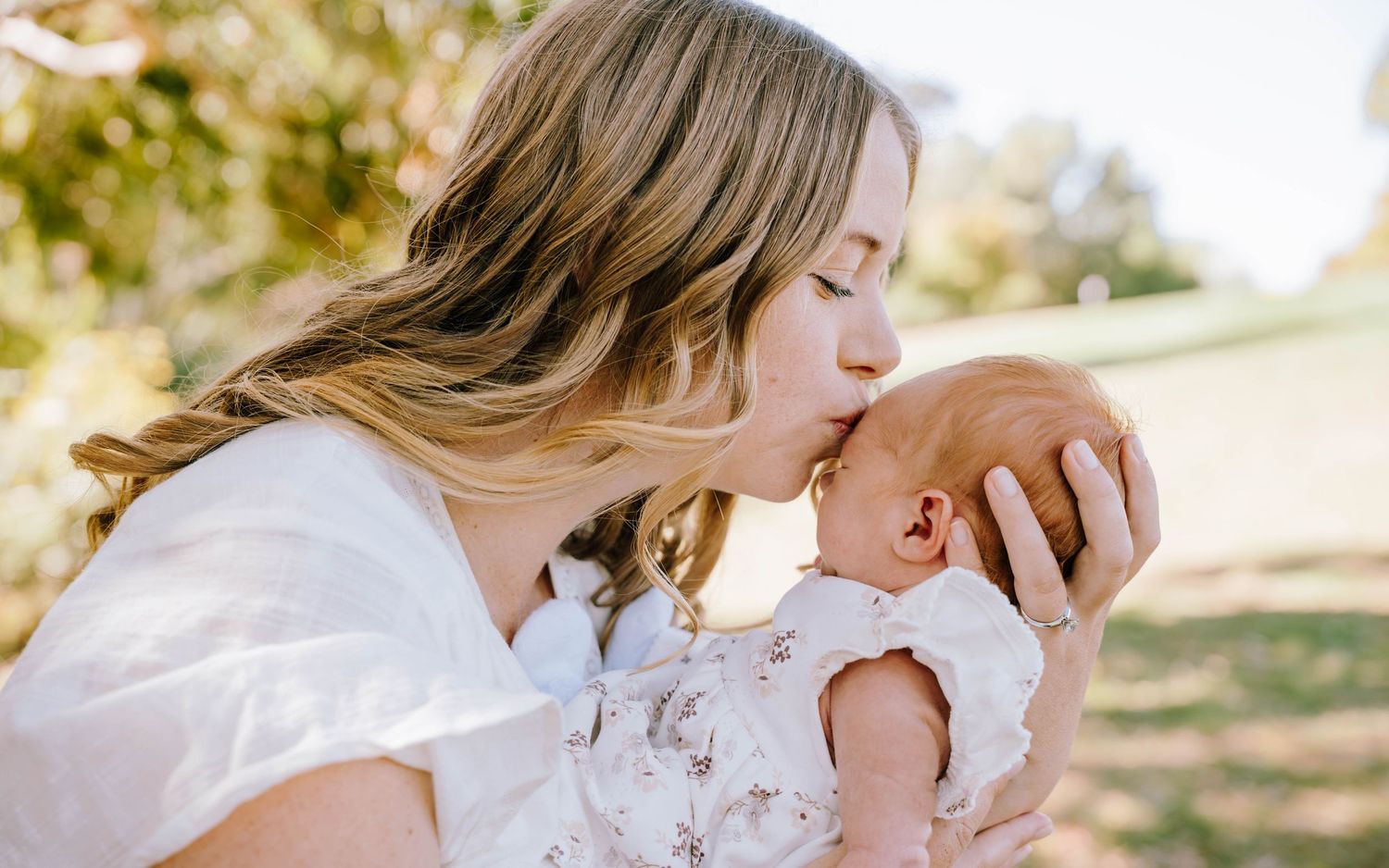 A mother tenderly kisses her newborn baby outdoors in a sunlit park setting.