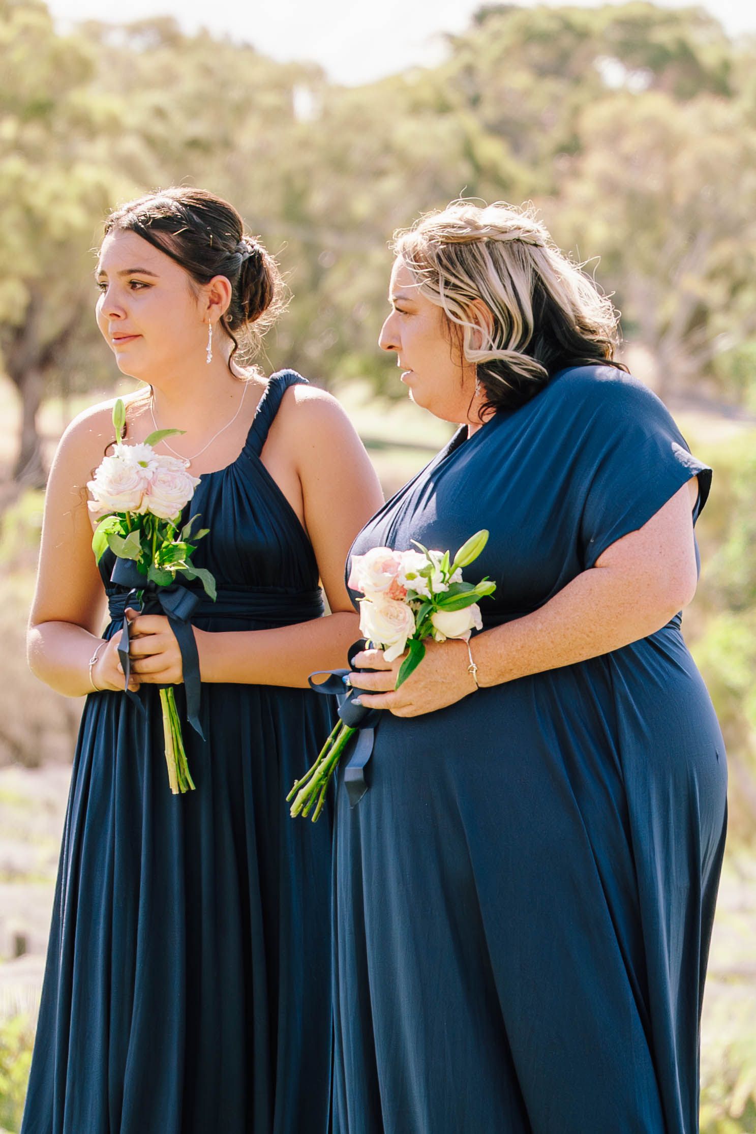 Two bridesmaids in navy blue dresses holding pink bouquets stand outdoors at a wedding ceremony.