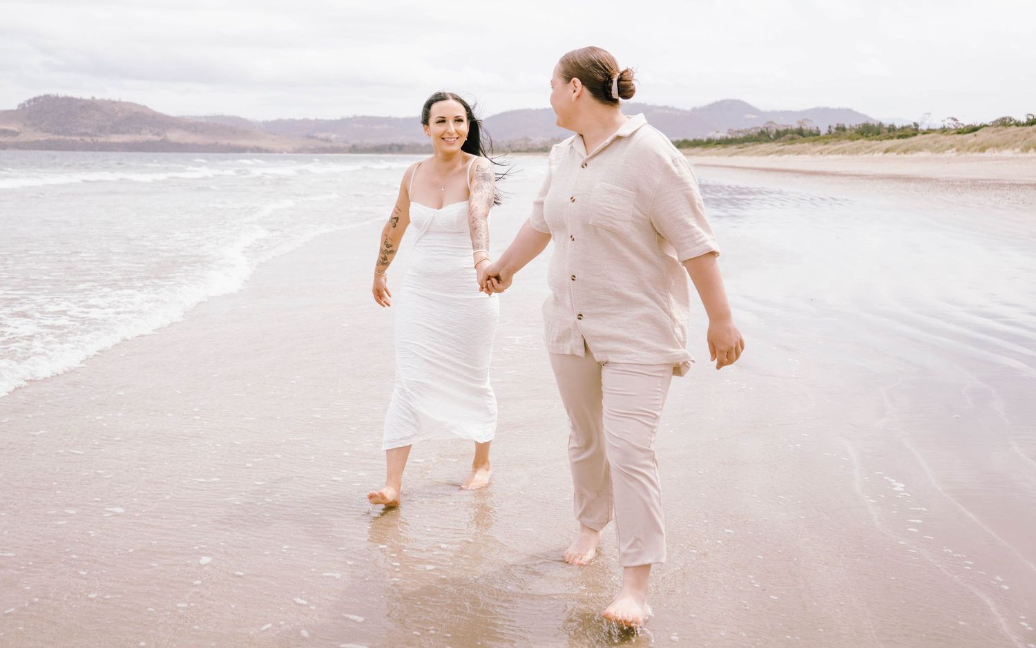 Couple walking hand in hand along a serene beach shoreline wearing white and beige outfits.