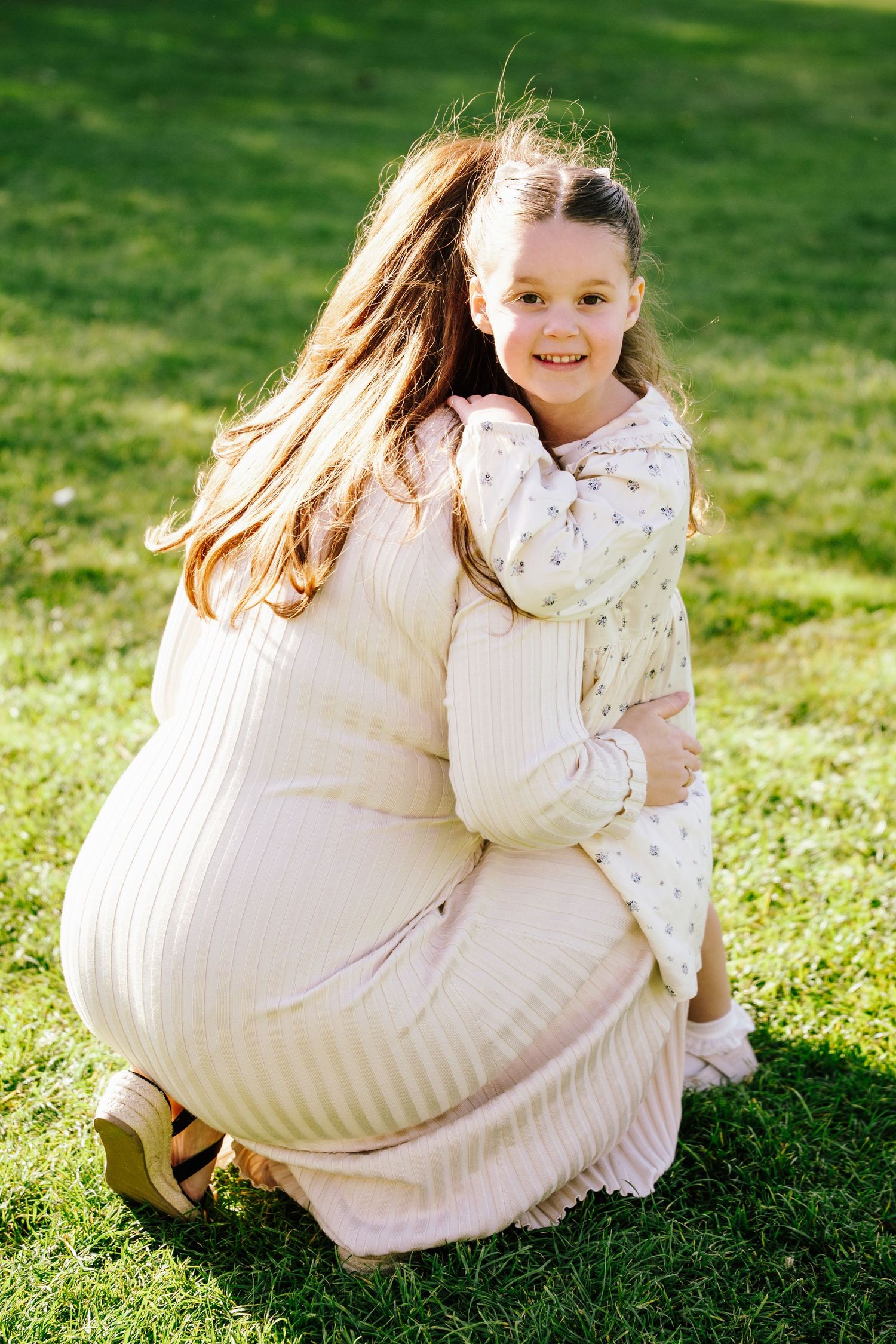Mother crouches to hug smiling young daughter in floral dress on green grass.